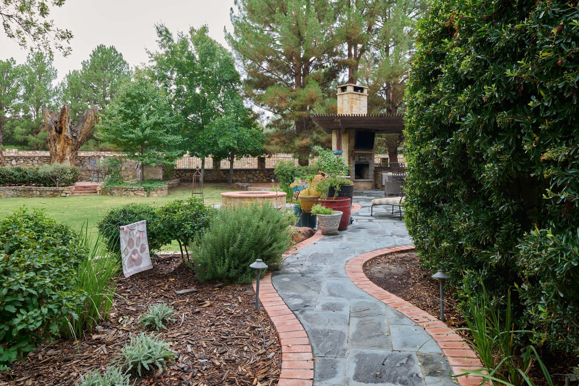 Stone path winds through a garden towards an outdoor fireplace; trees and shrubs border the path.