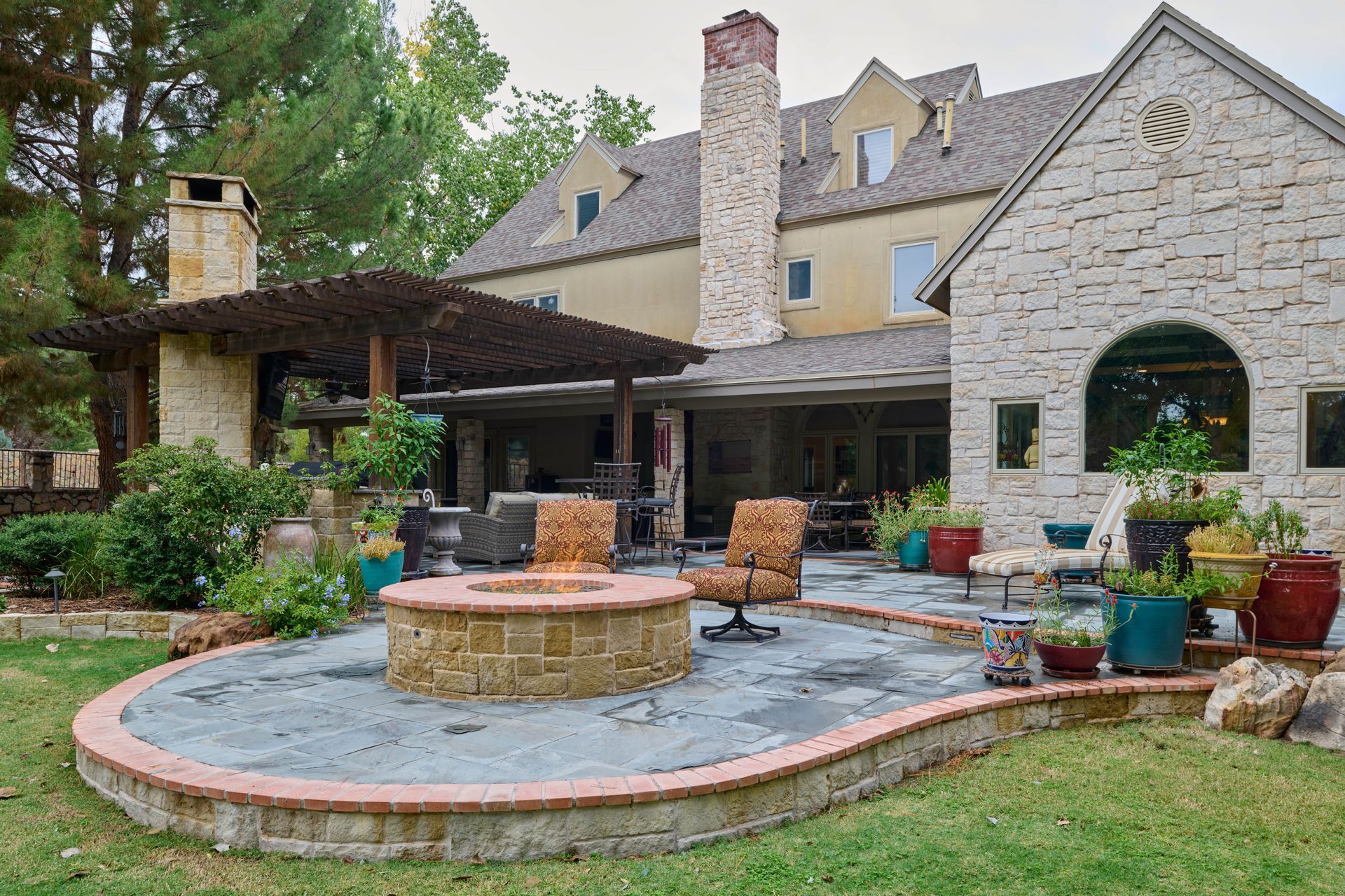Backyard patio with fire pit, seating, and multi-story stone house. Green lawn, brick, and gray pavers.