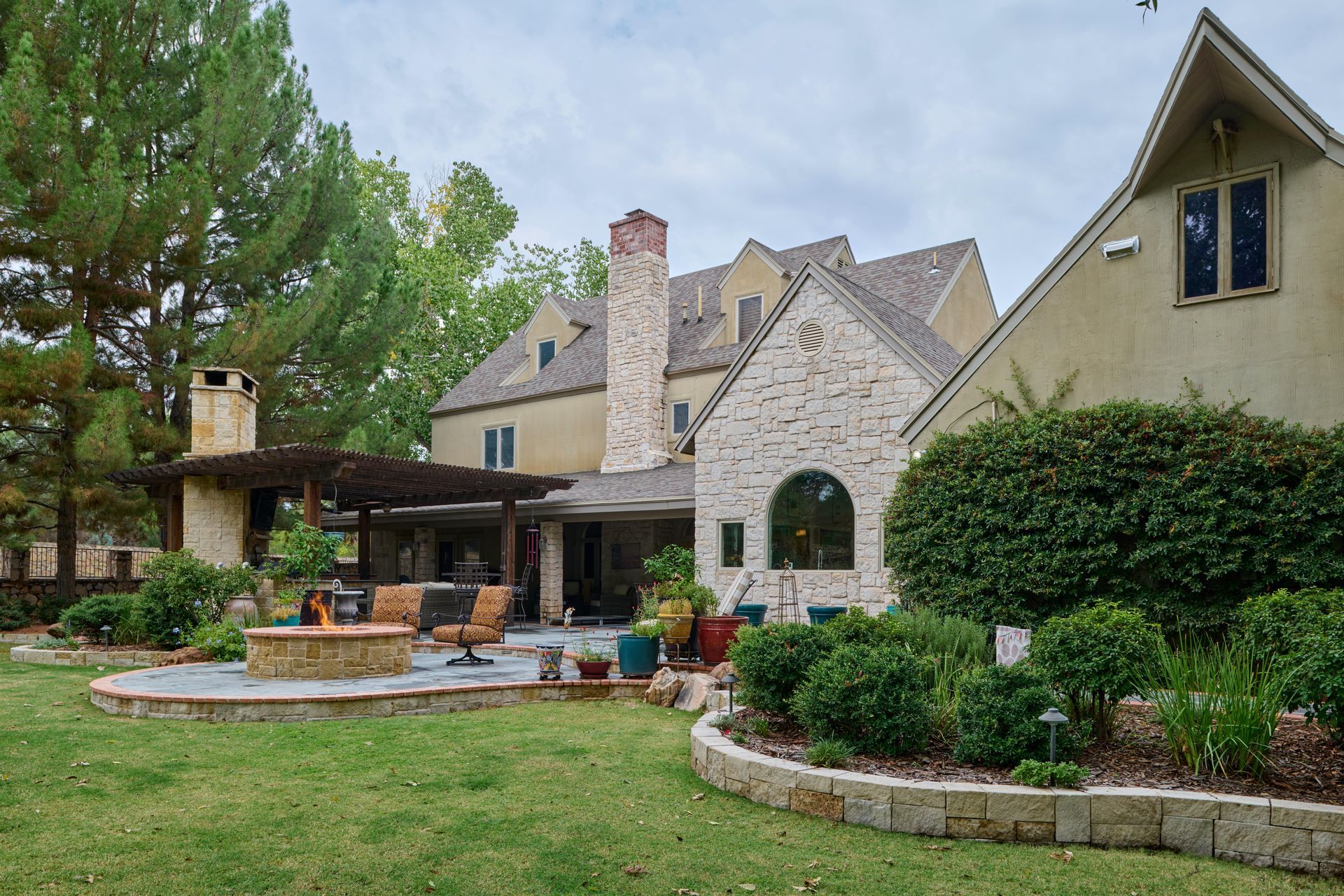 Backyard with house, patio, fire pit, and landscaping under cloudy sky.