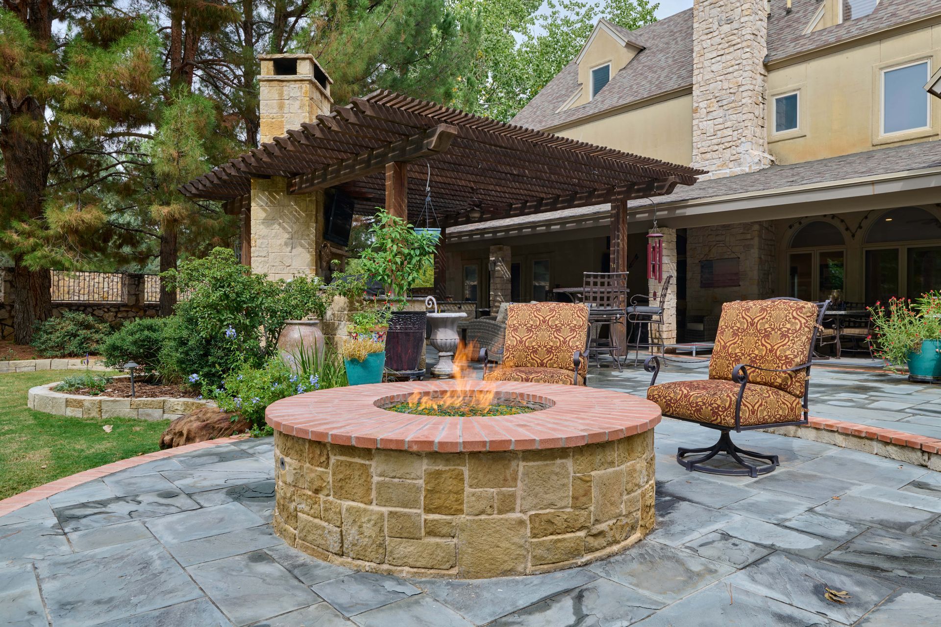 Stone patio with a fire pit, seating, and a house in the background. Fire is lit.