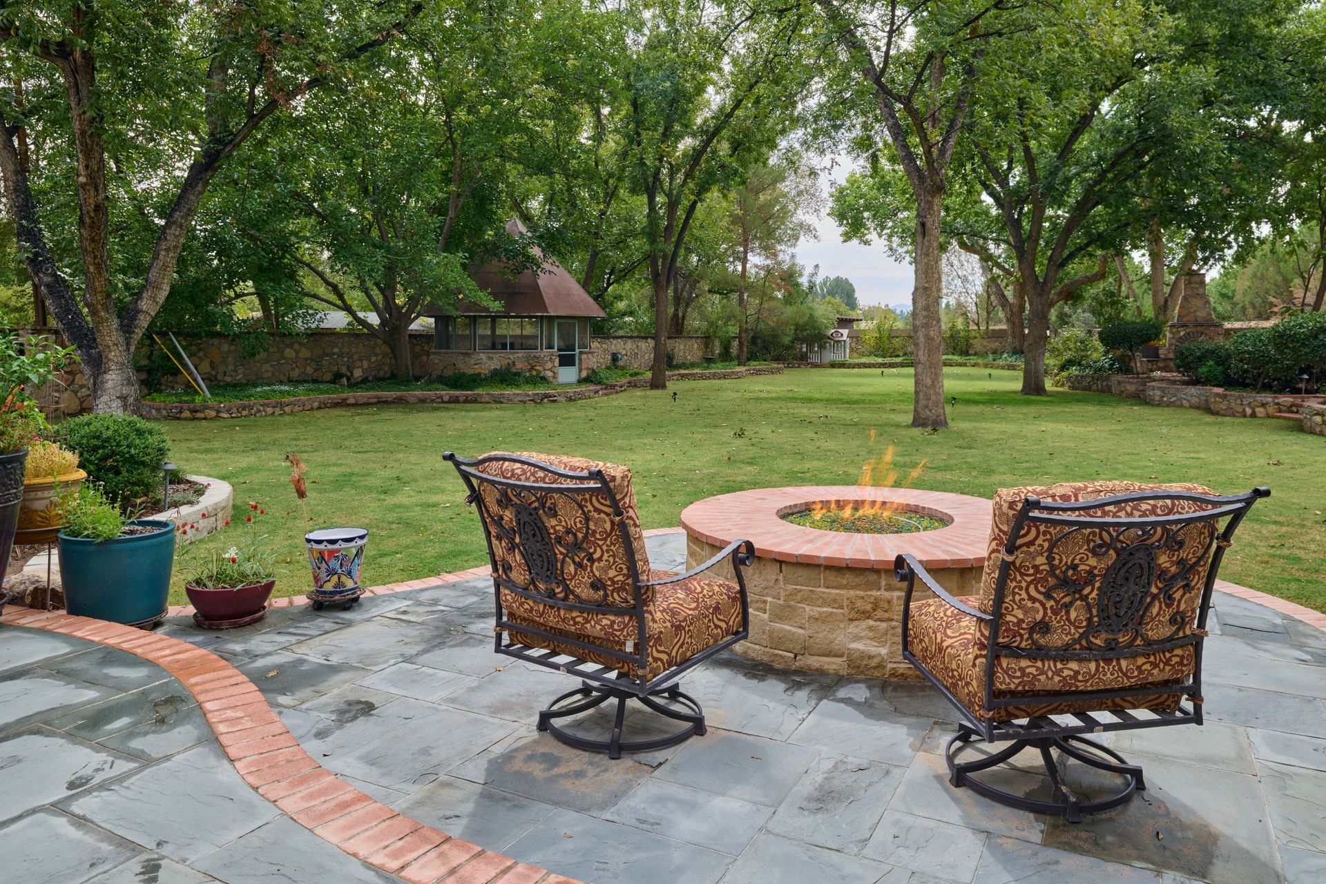 Two chairs face a fire pit on a patio, overlooking a grassy backyard with trees.