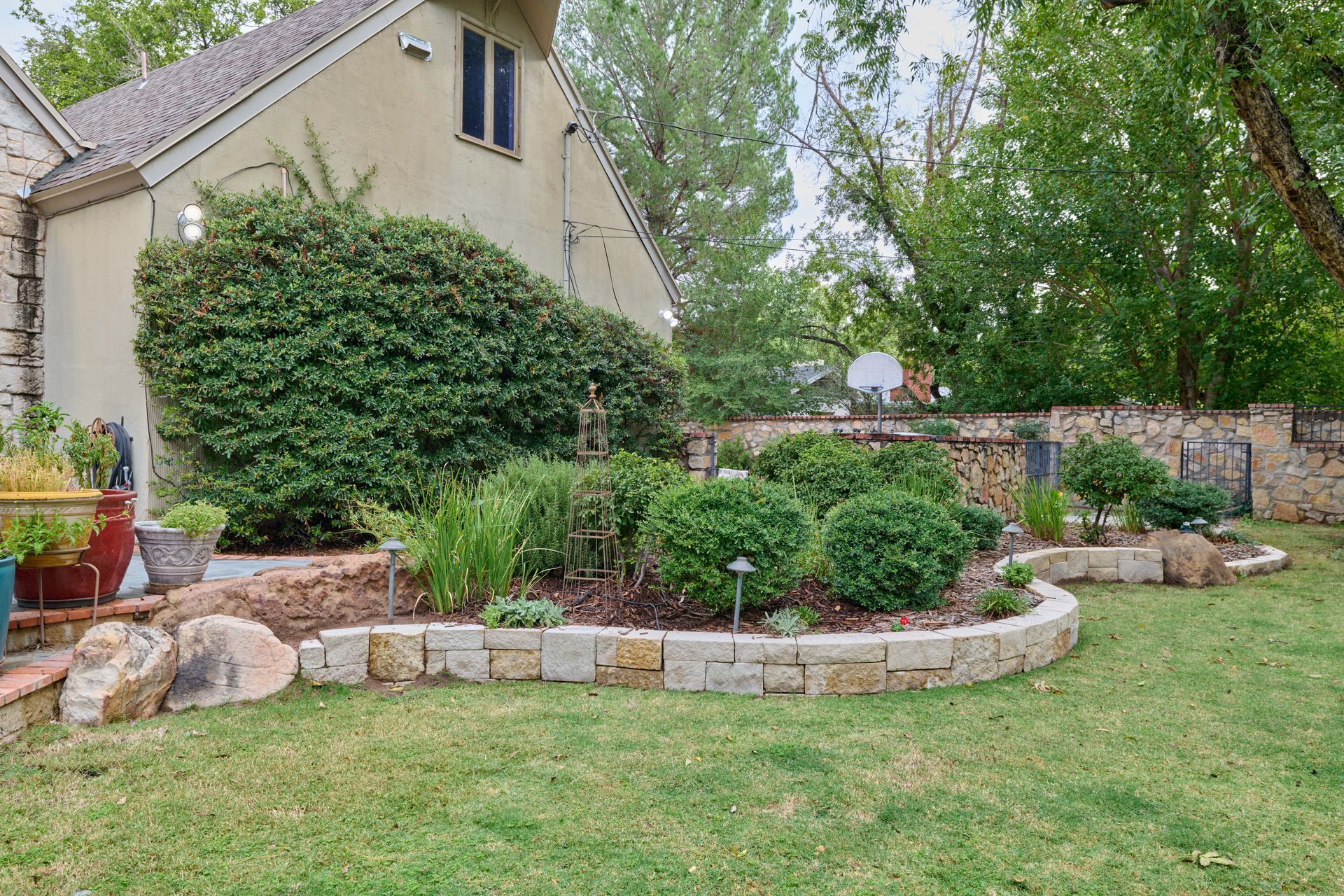 Backyard garden with stone-edged flower beds and lush greenery against a beige house.