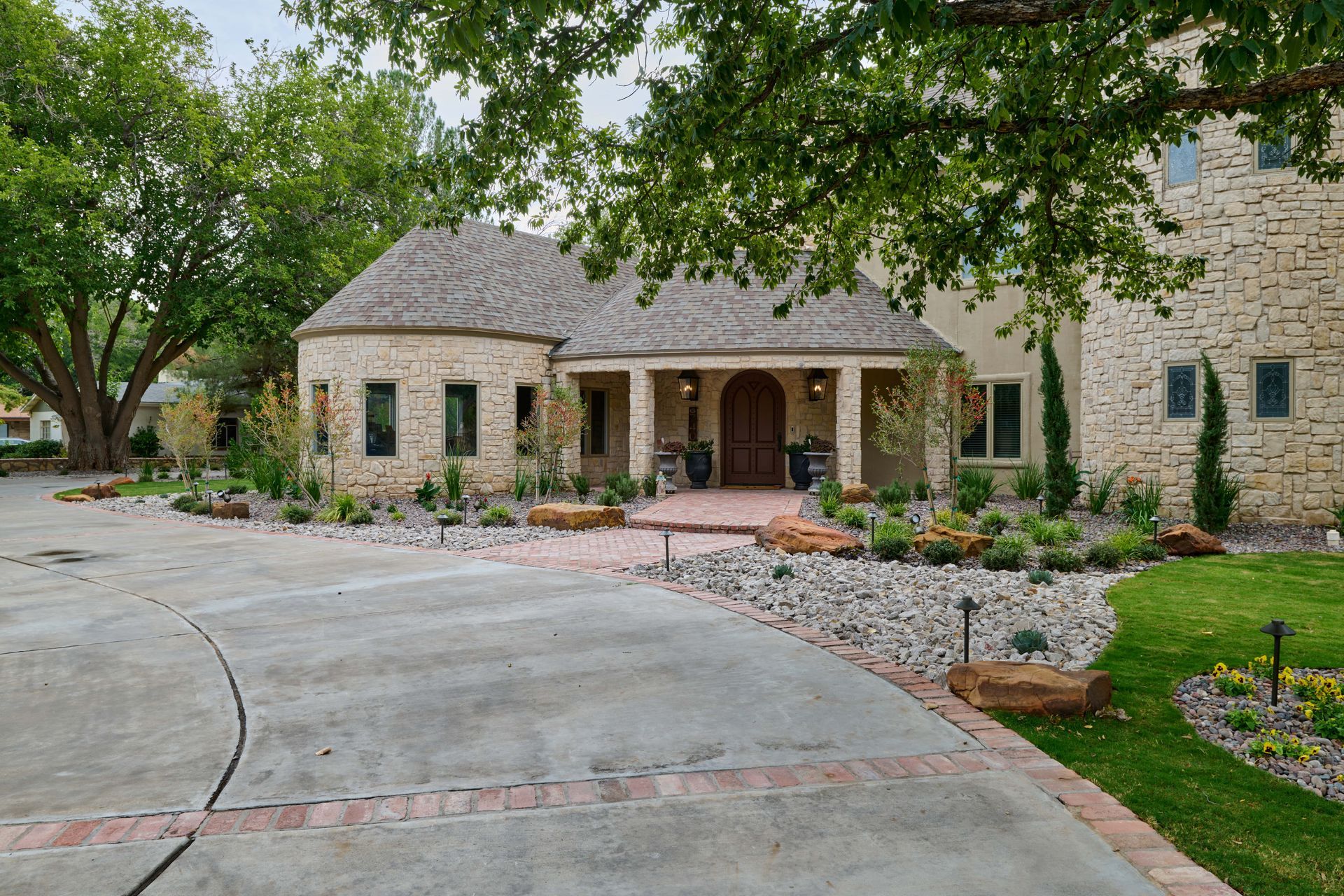 Stone-faced house with a circular driveway and manicured landscaping under a cloudy sky.