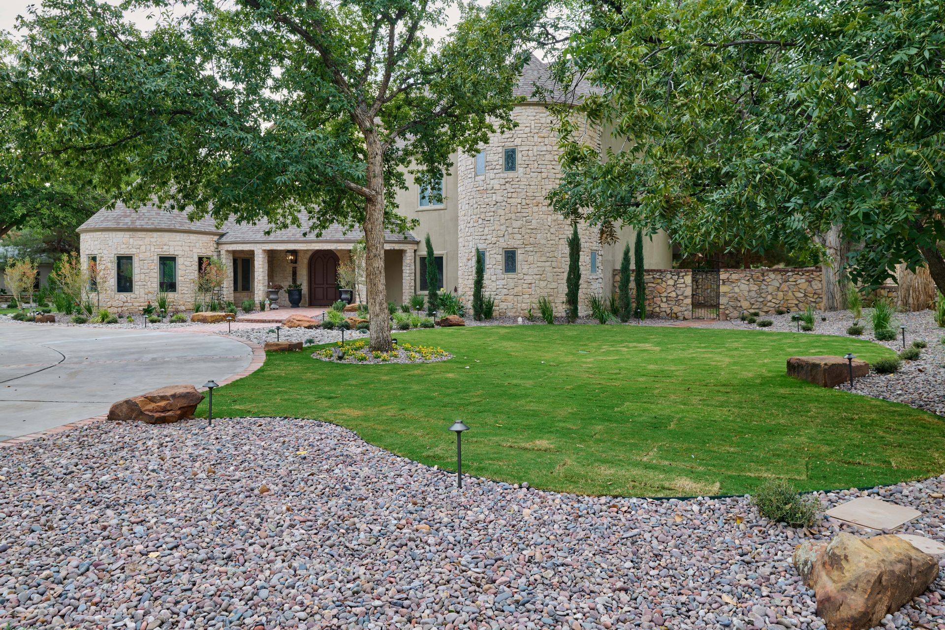 Stone house with green lawn, gravel drive, and trees.