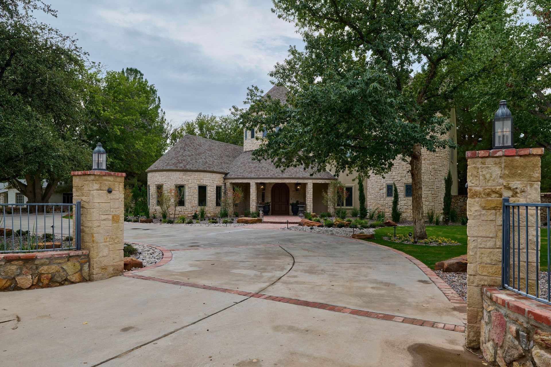 Stone house with long driveway, framed by pillars with lamps and lush greenery. Overcast sky.