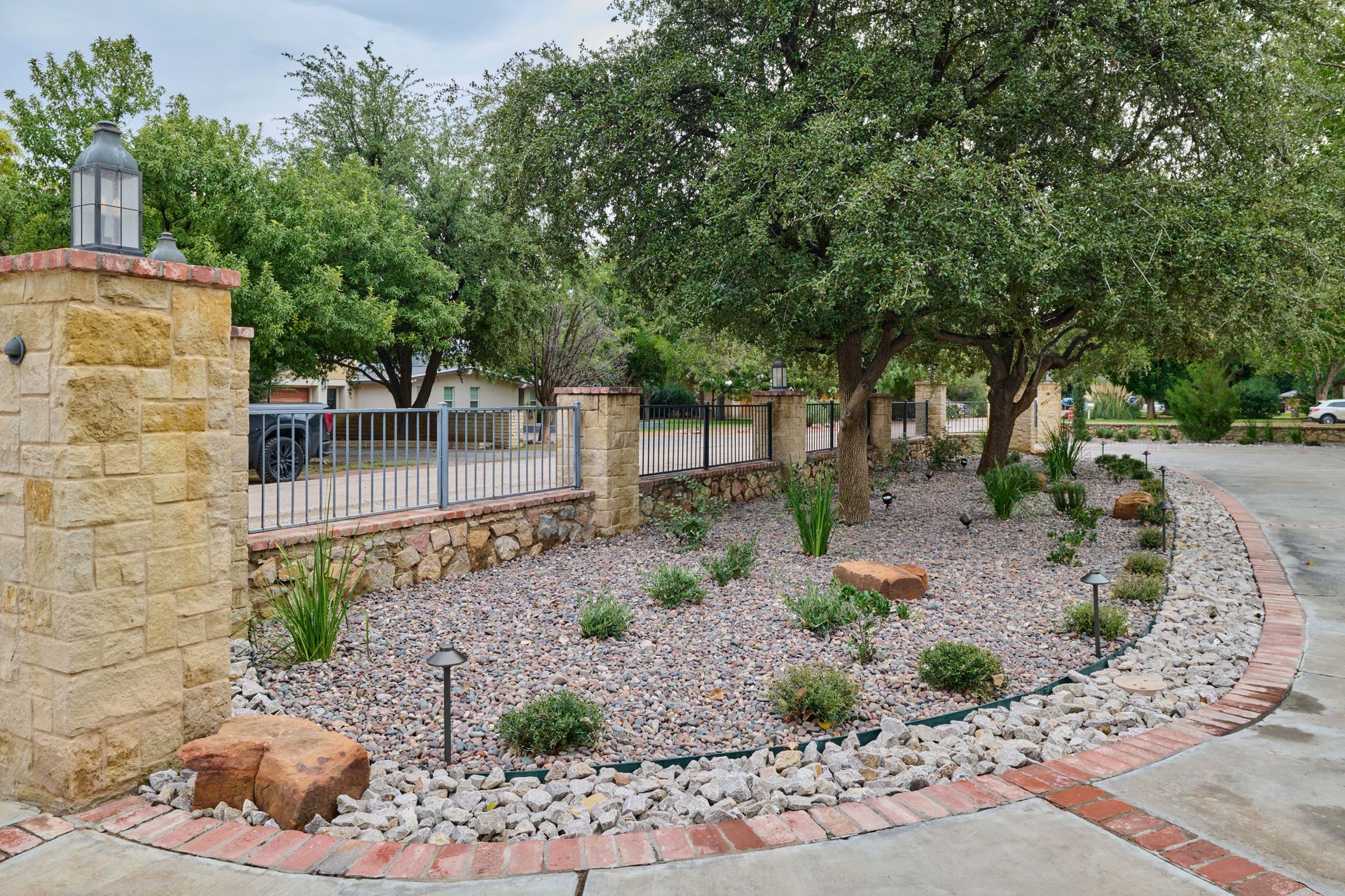 Stone pillars frame a landscaped bed with gravel, small plants, and trees alongside a brick-lined driveway.