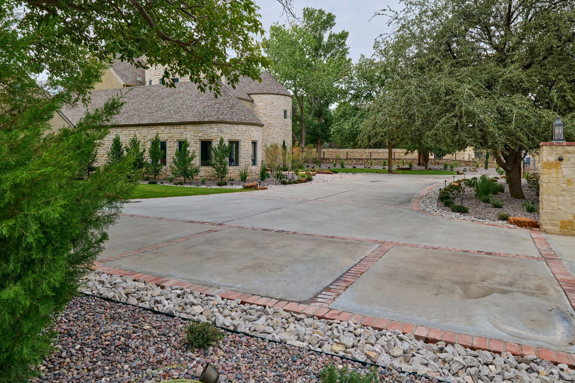 Driveway leading to a stone house with a round turret, surrounded by trees and landscaping.