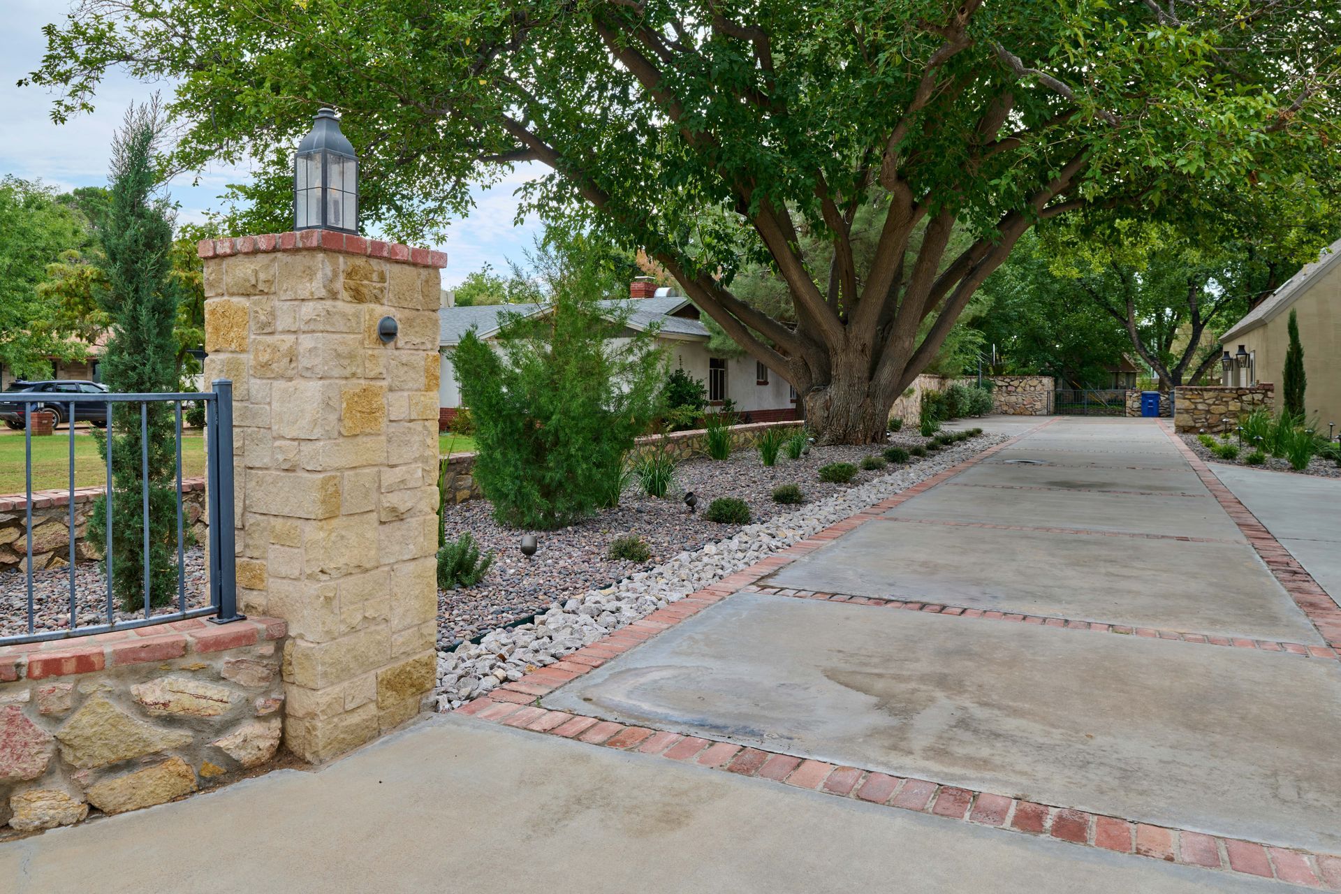 Driveway entrance with stone pillars, brick borders, and large tree.