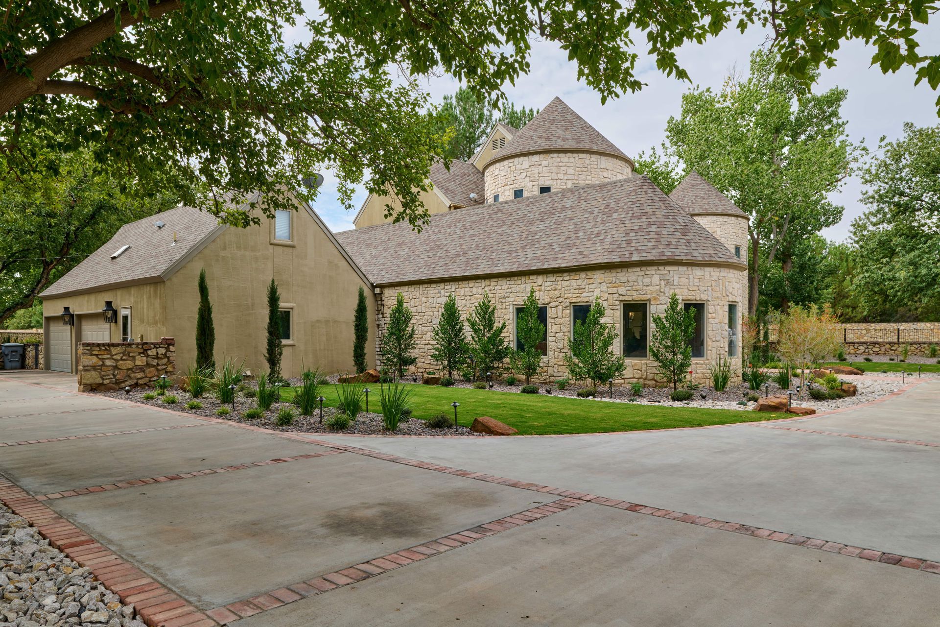 Stone and stucco house with a circular turret, nestled under trees. Driveway in foreground.