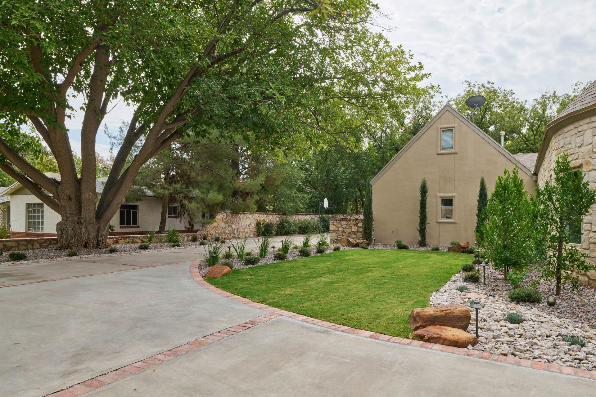 Driveway and lawn of a house with trees and a light-colored building.