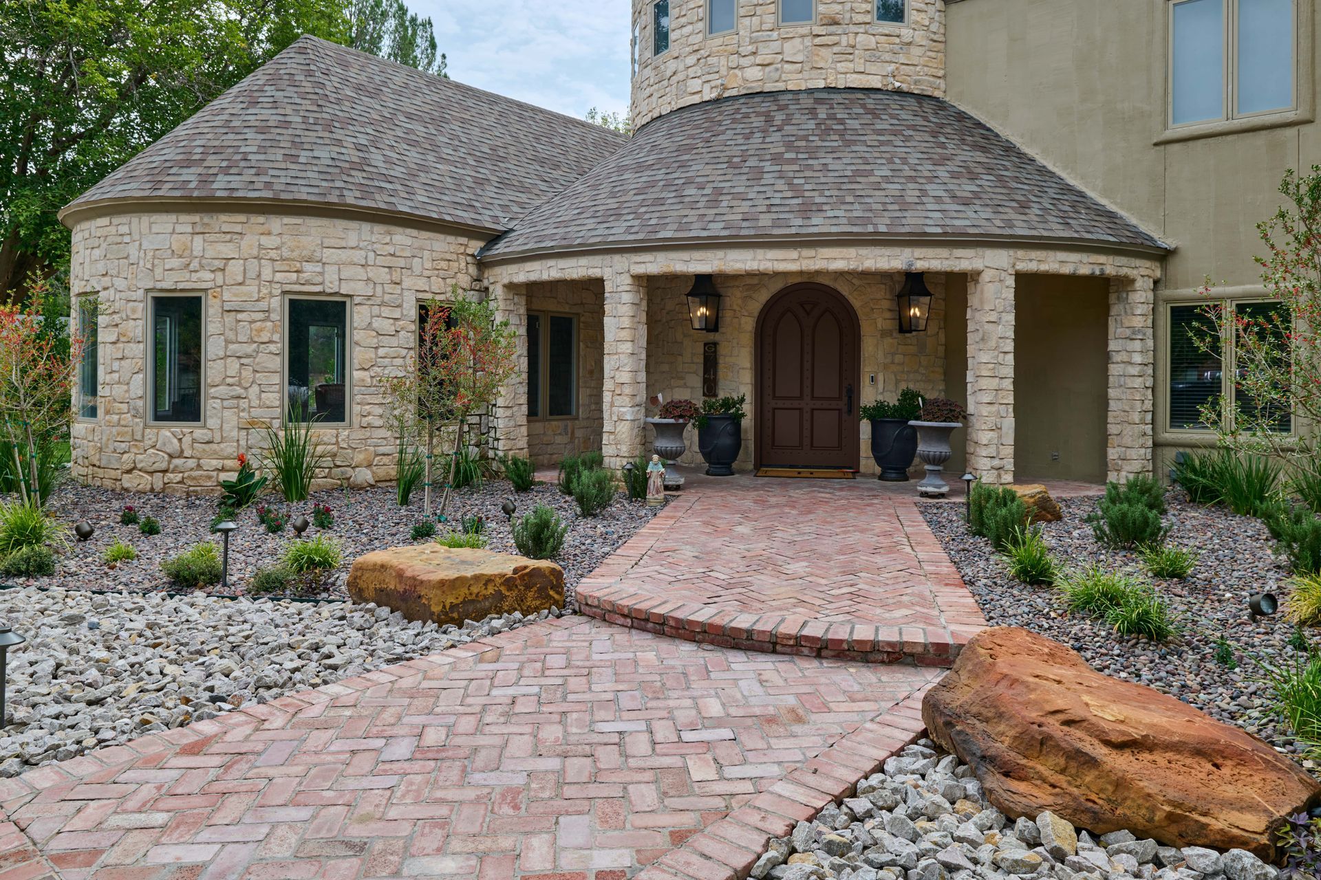 Brick pathway leading to a tan stone house with a curved roof and arched entrance. Landscaping with rocks and plants.
