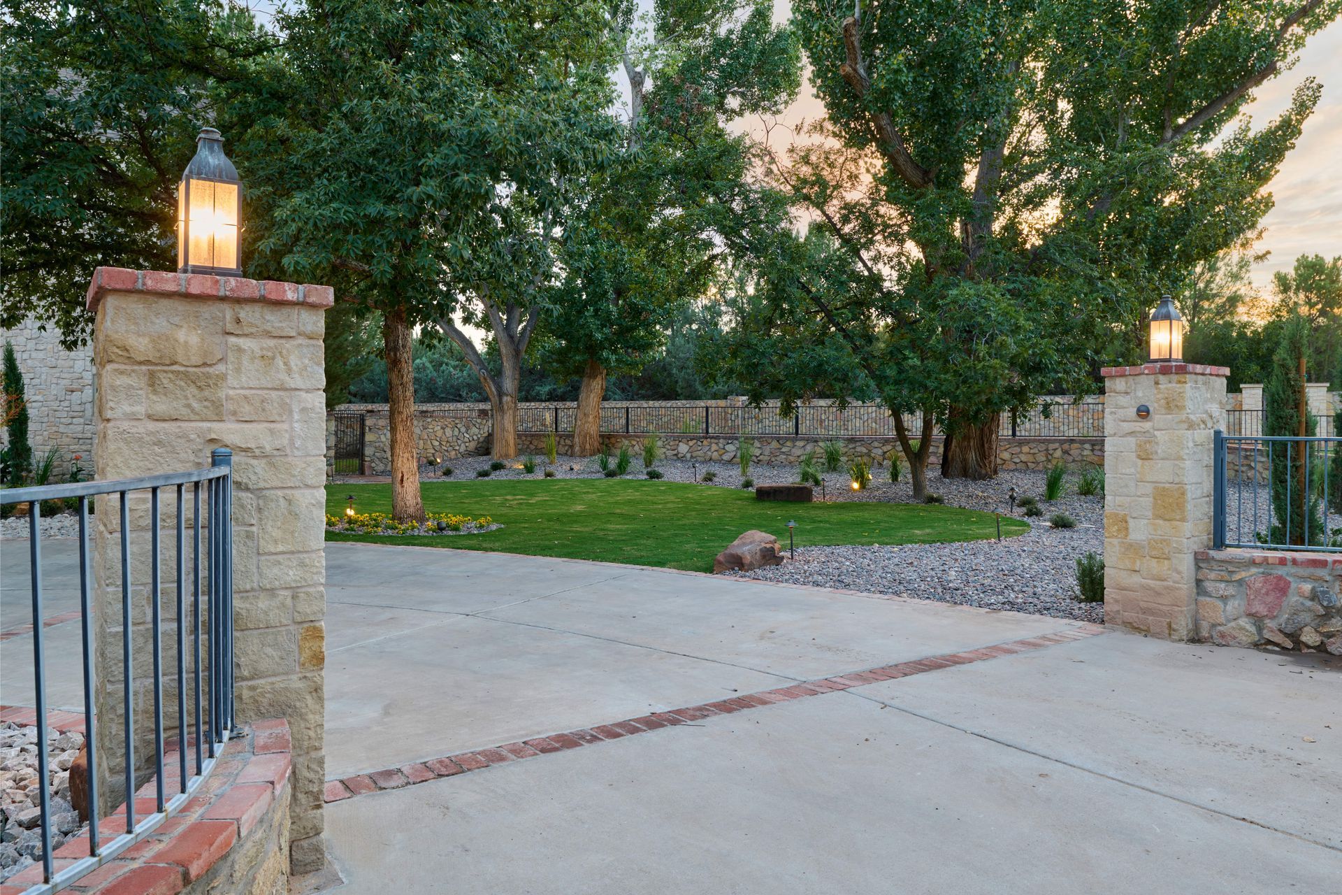 Entryway with stone pillars, lanterns, concrete drive, lawn, trees, and a low stone wall.