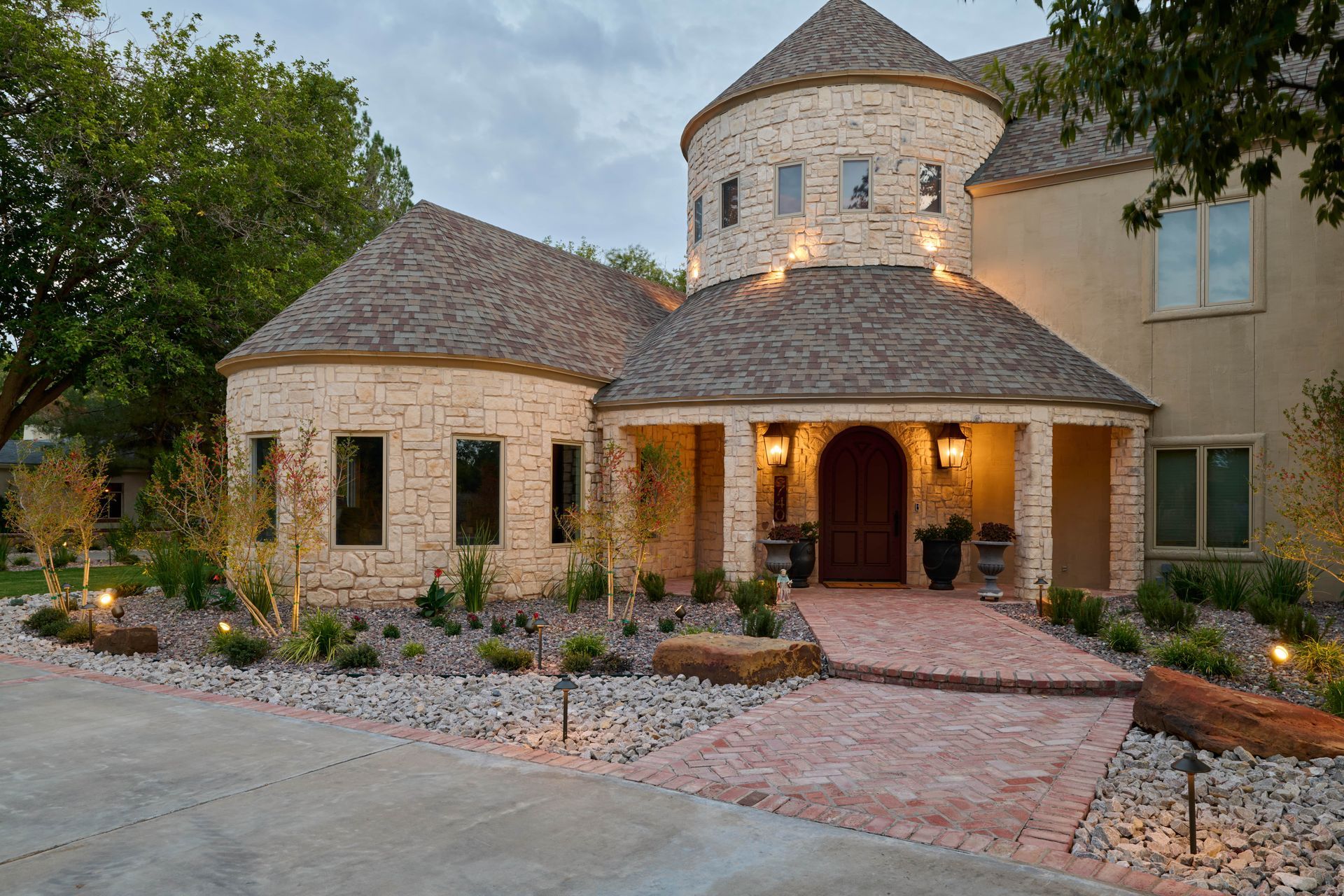 Stone house with a circular turret entrance, brick pathway, and landscape lighting at dusk.