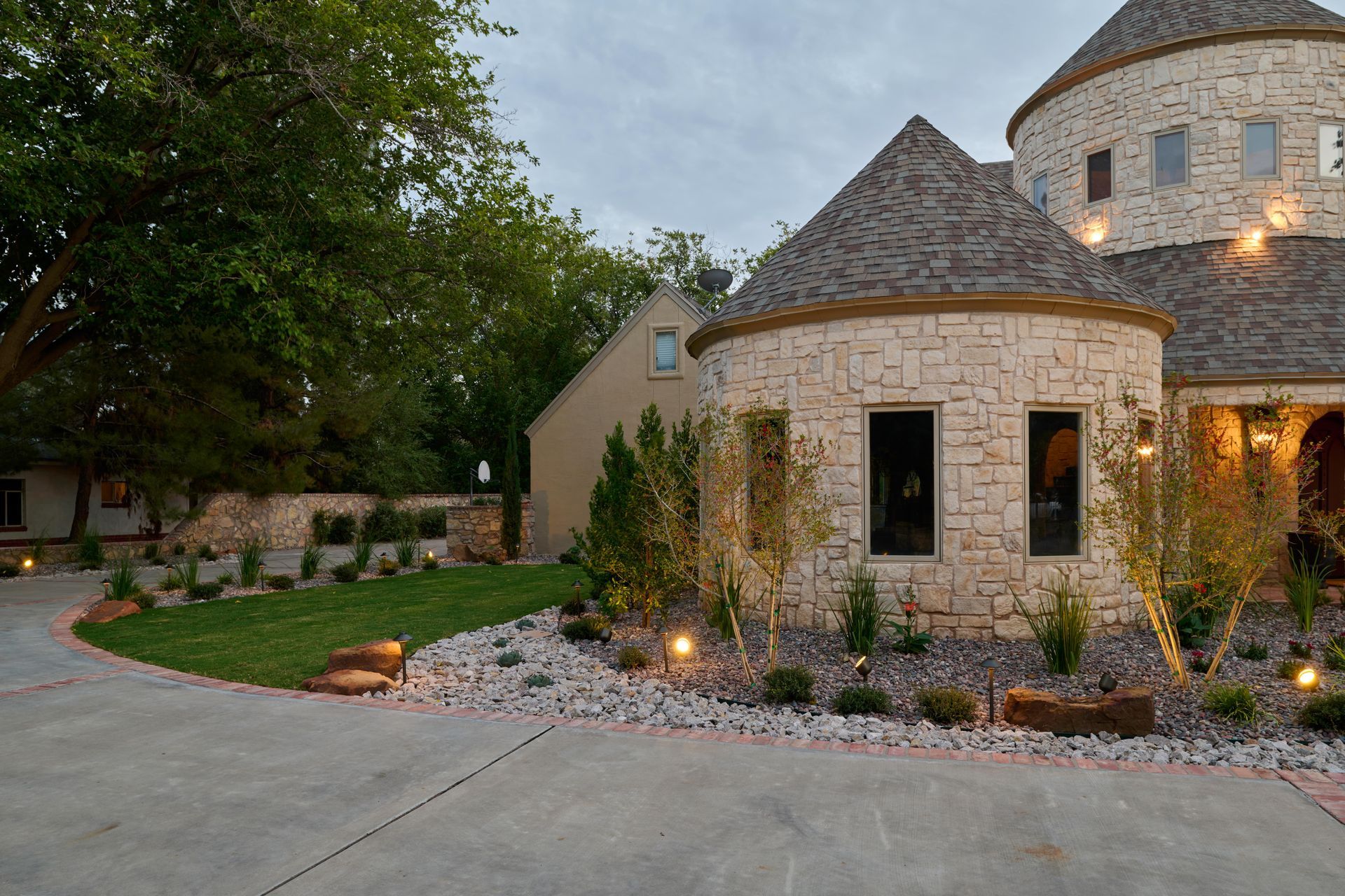 Stone house with turrets and a circular driveway lit by landscape lights.