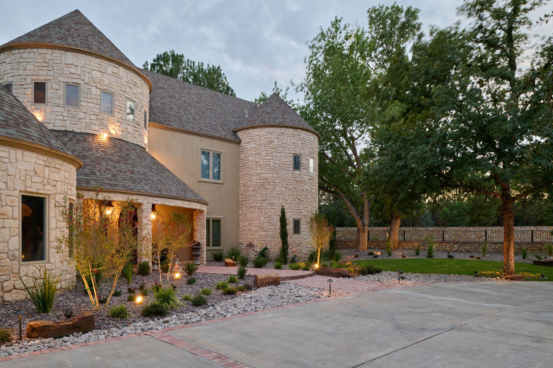 Stone castle-style house with round turrets, concrete driveway, and landscaped yard at dusk.