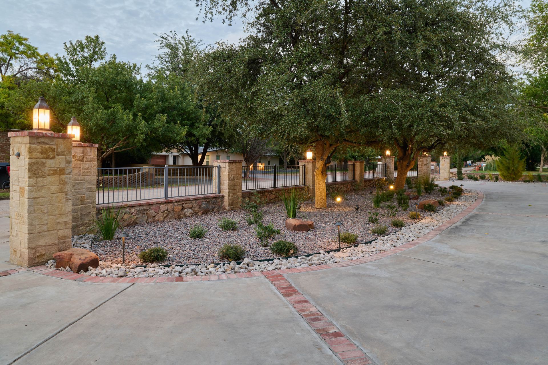 Entrance with stone pillars, trees, lighting, and a brick-lined driveway.