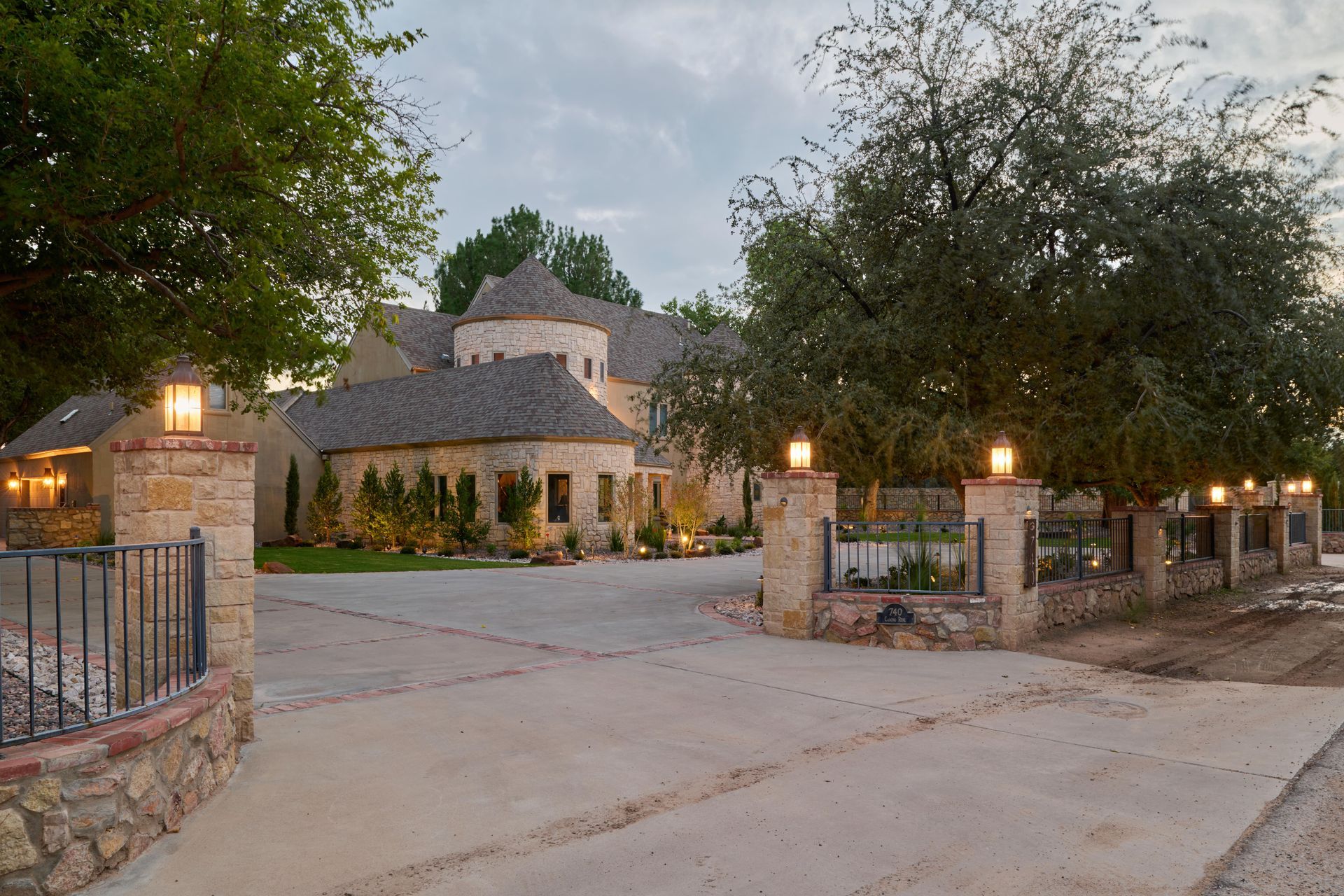 Stone house with a circular turret, accessed by a concrete driveway lined with lighted posts and a stone wall.