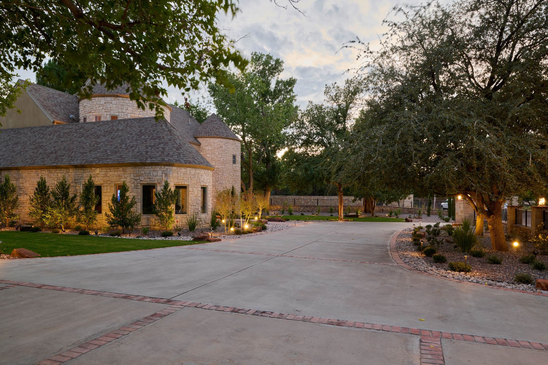 Stone castle-like building with driveway, surrounded by trees and landscaping, illuminated at dusk.