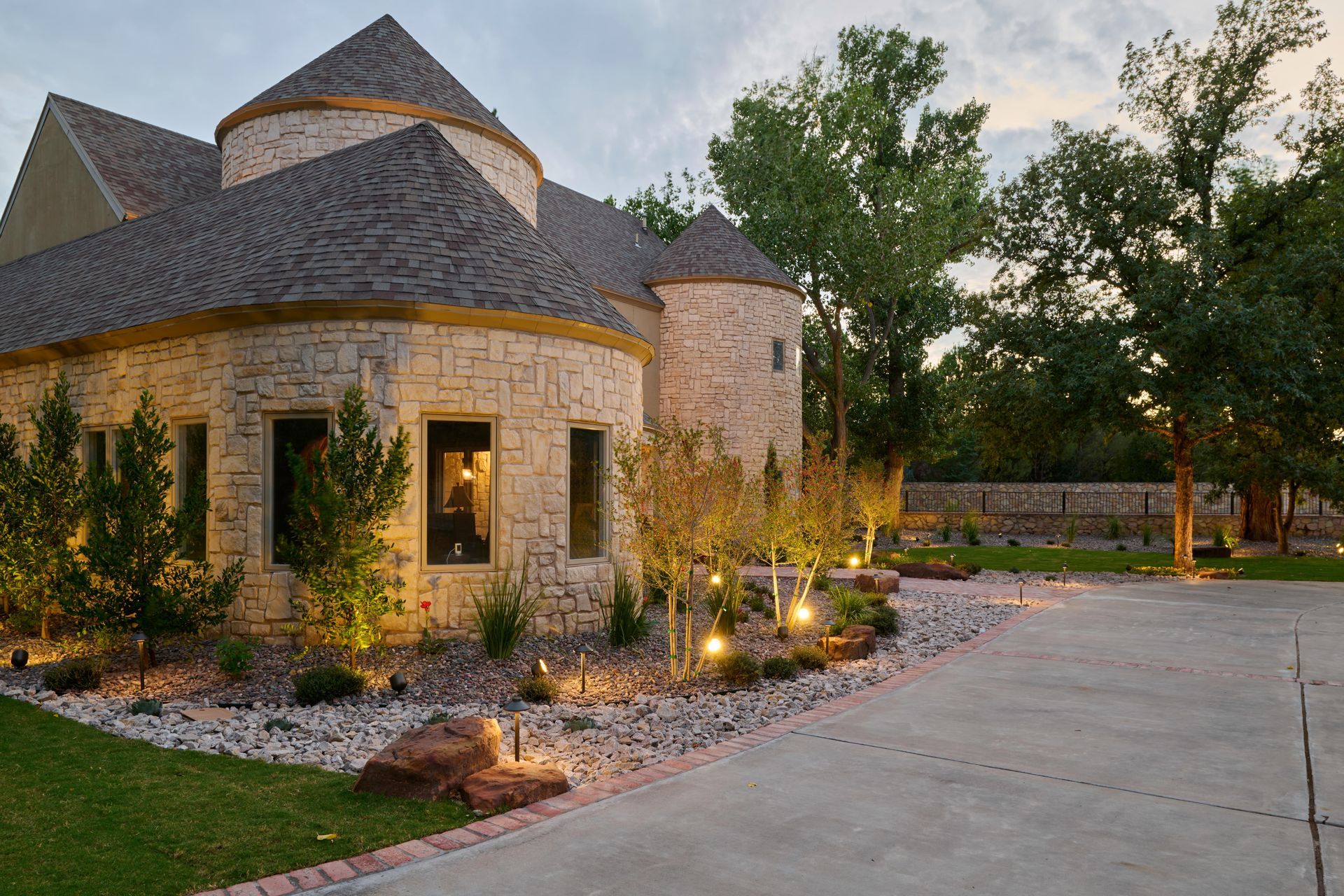 Elegant stone house with turret, illuminated landscaping, and long driveway.
