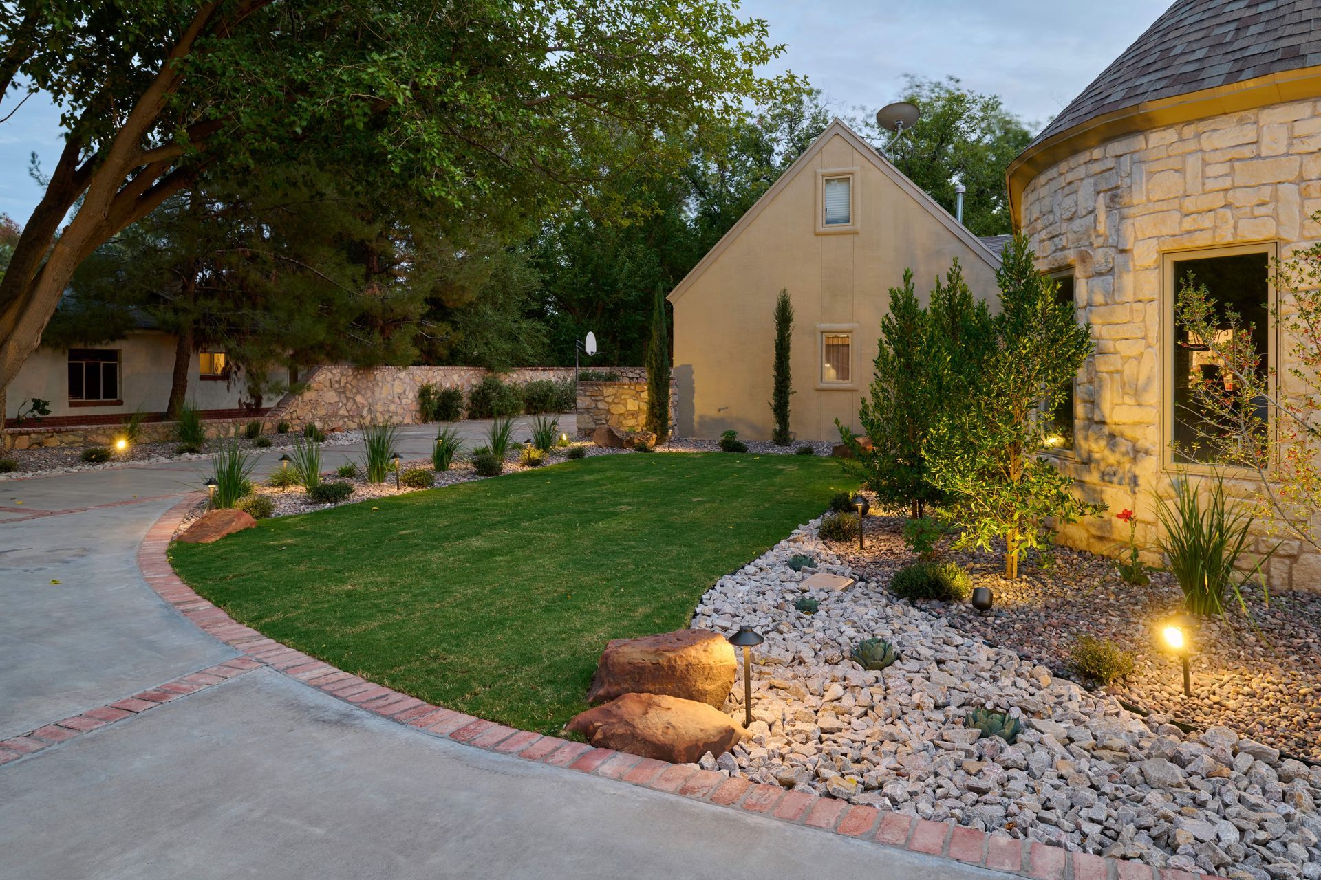 Curving driveway leads to a stone house and beige outbuilding, manicured lawn, and decorative rock garden.