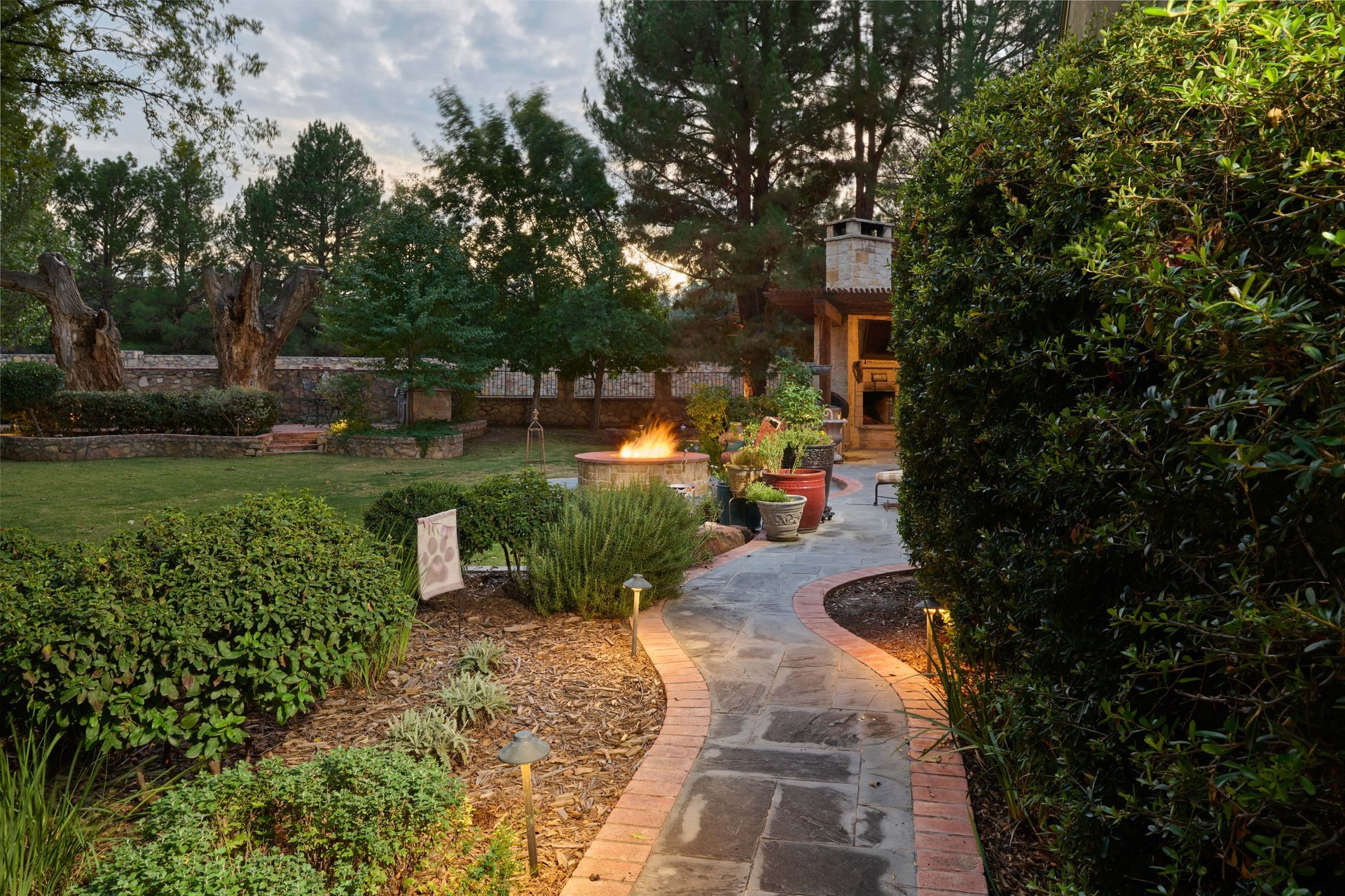 Stone path in a garden with a fire pit, lined with bushes, and lights illuminating the way.