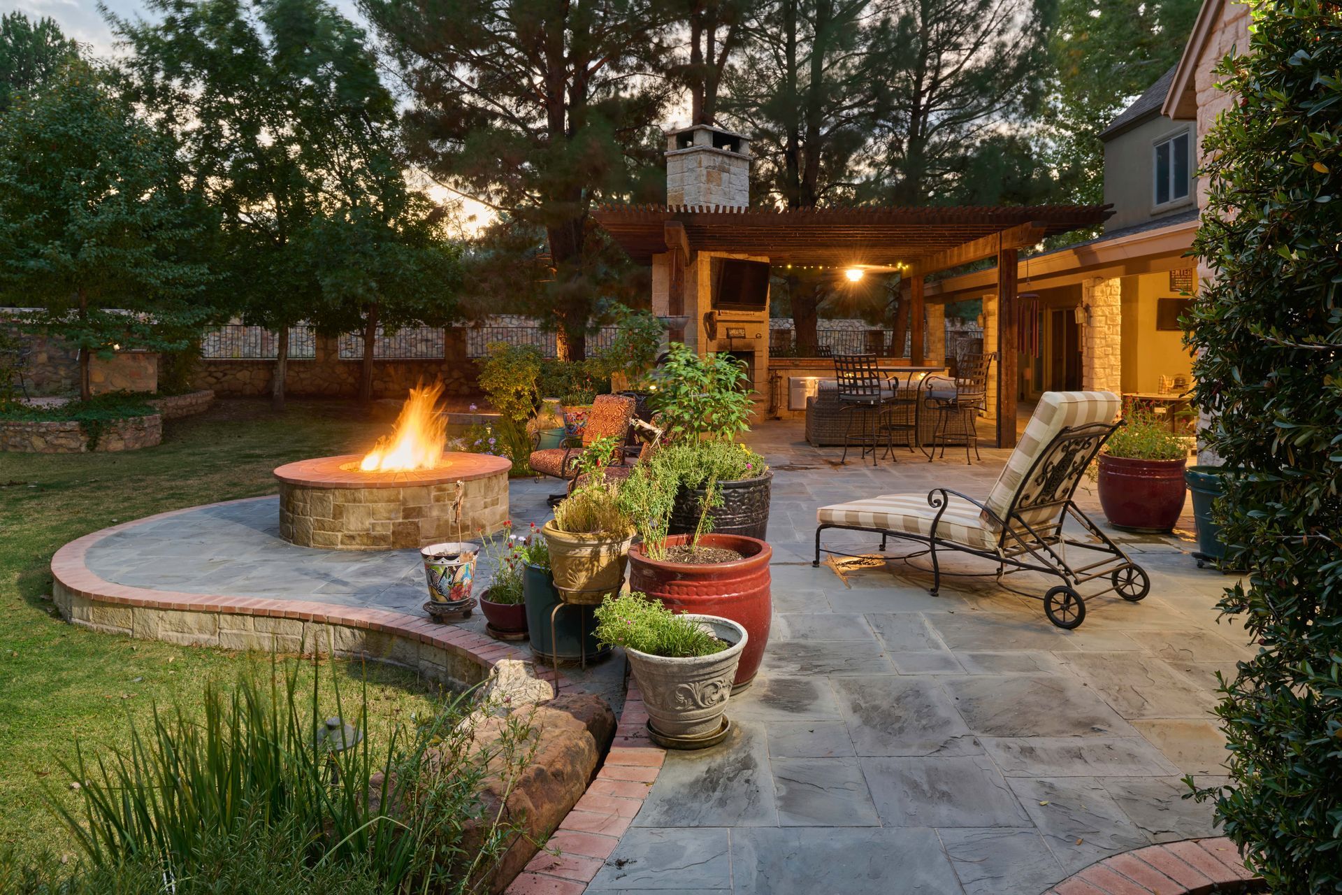 Backyard patio with fire pit, lounge chairs, pergola, and plants at dusk.