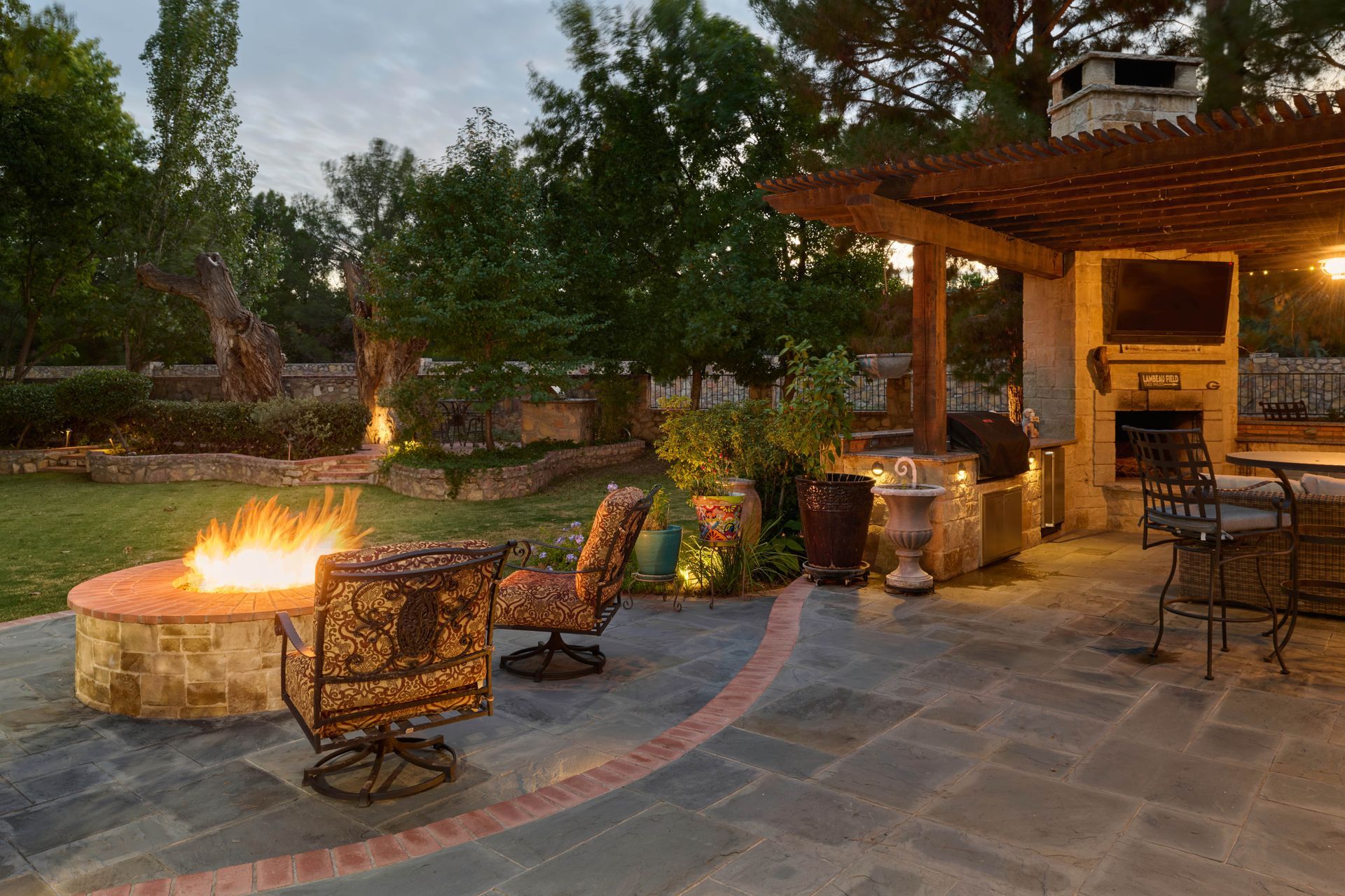 Stone patio with fire pit and outdoor seating, trees in the background, lit at dusk.