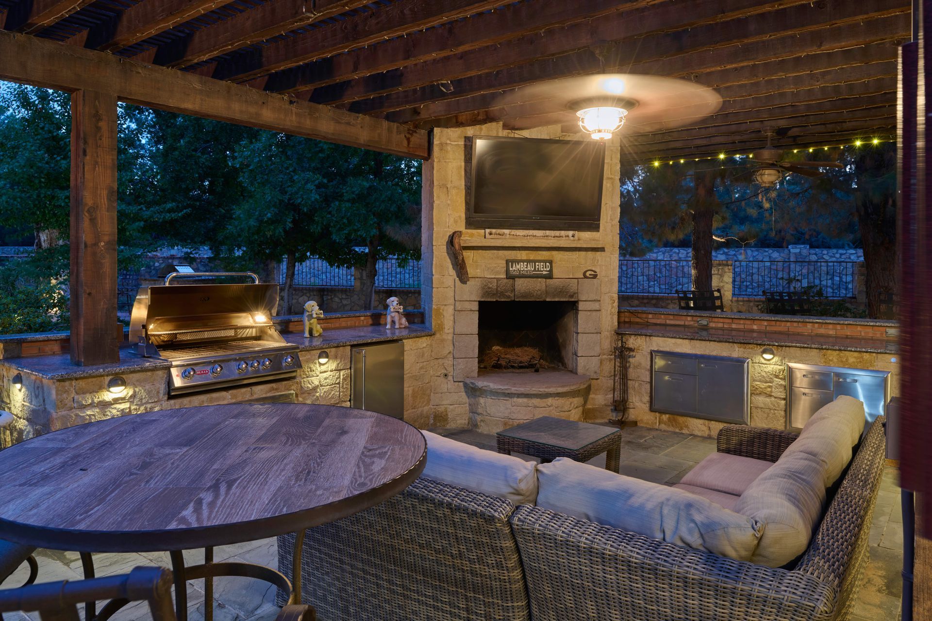 Outdoor kitchen with grill, fireplace, TV, seating, and wooden table under a pergola at dusk.