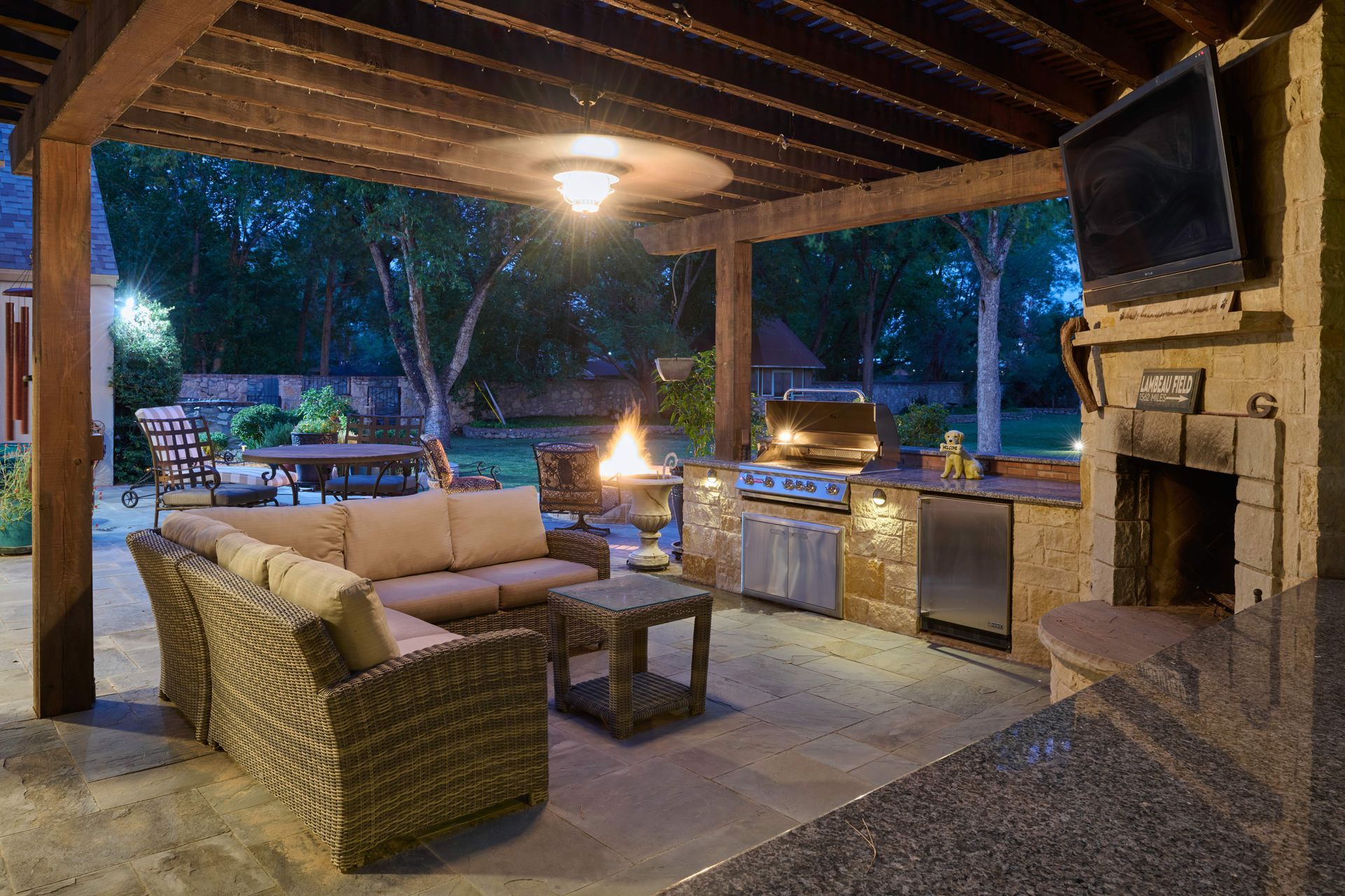 Outdoor kitchen and lounge with fire pit, grill, TV, seating, and wooden pergola.