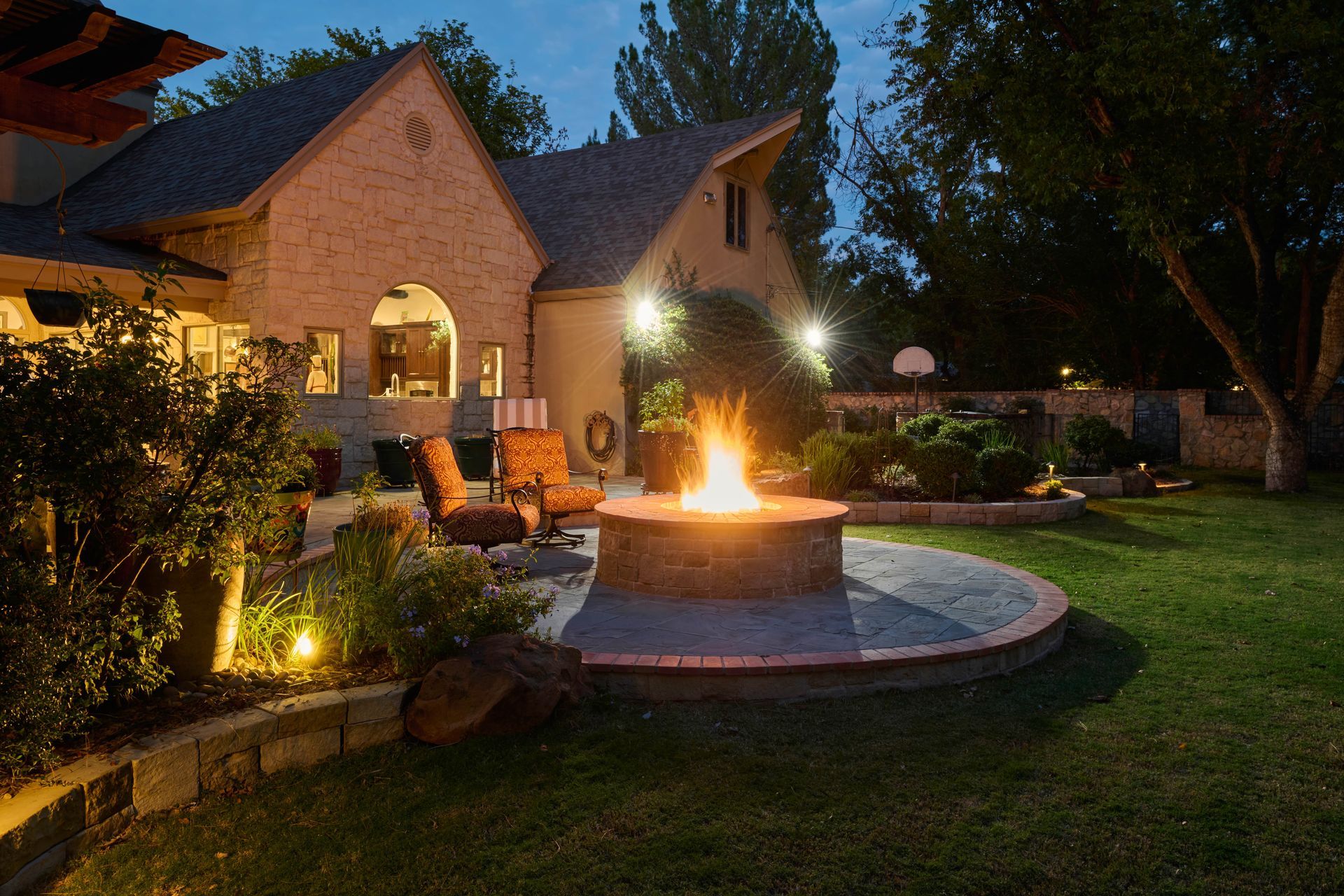 Backyard with fire pit, seating, and house at night, lit by spotlights.