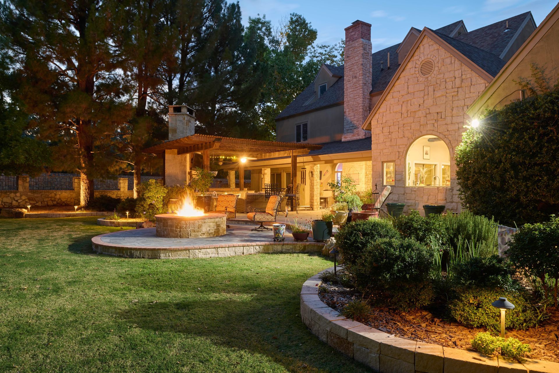 Backyard with fire pit, patio, and home at dusk; lush green lawn and landscape lighting.