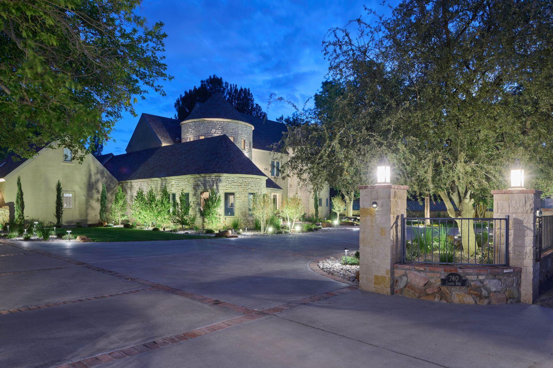 Stone house at dusk, illuminated driveway with gate, surrounded by trees.