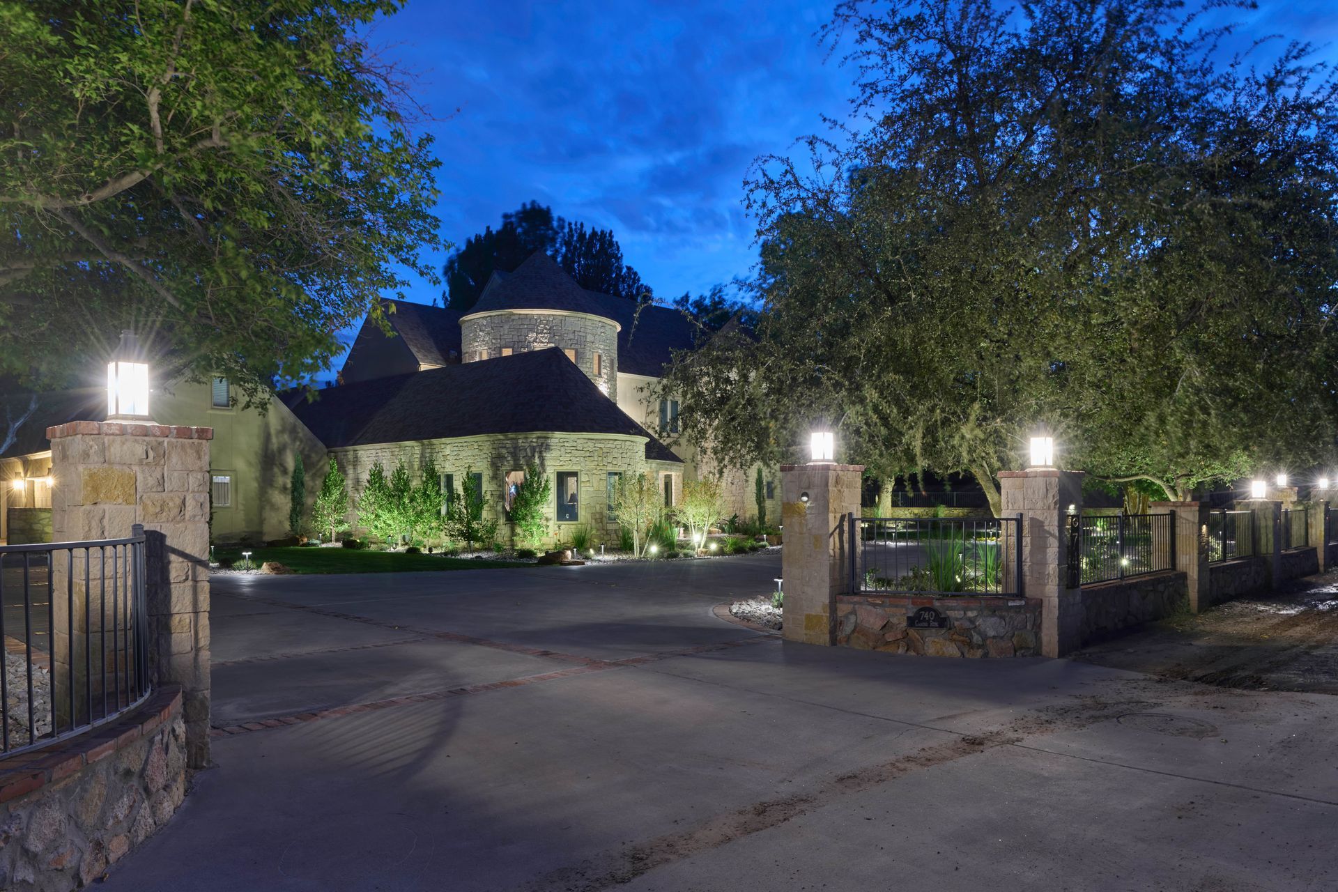 Stone house and driveway lit at dusk, with trees and outdoor lights.