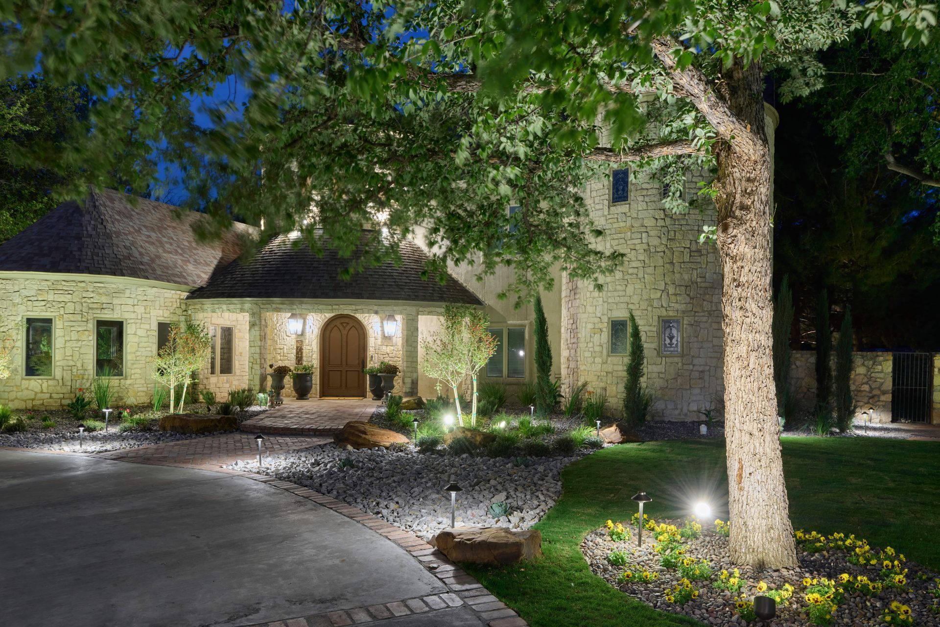 Lit stone house with arched doorway, tree, and landscaped yard. Driveway and pathway.
