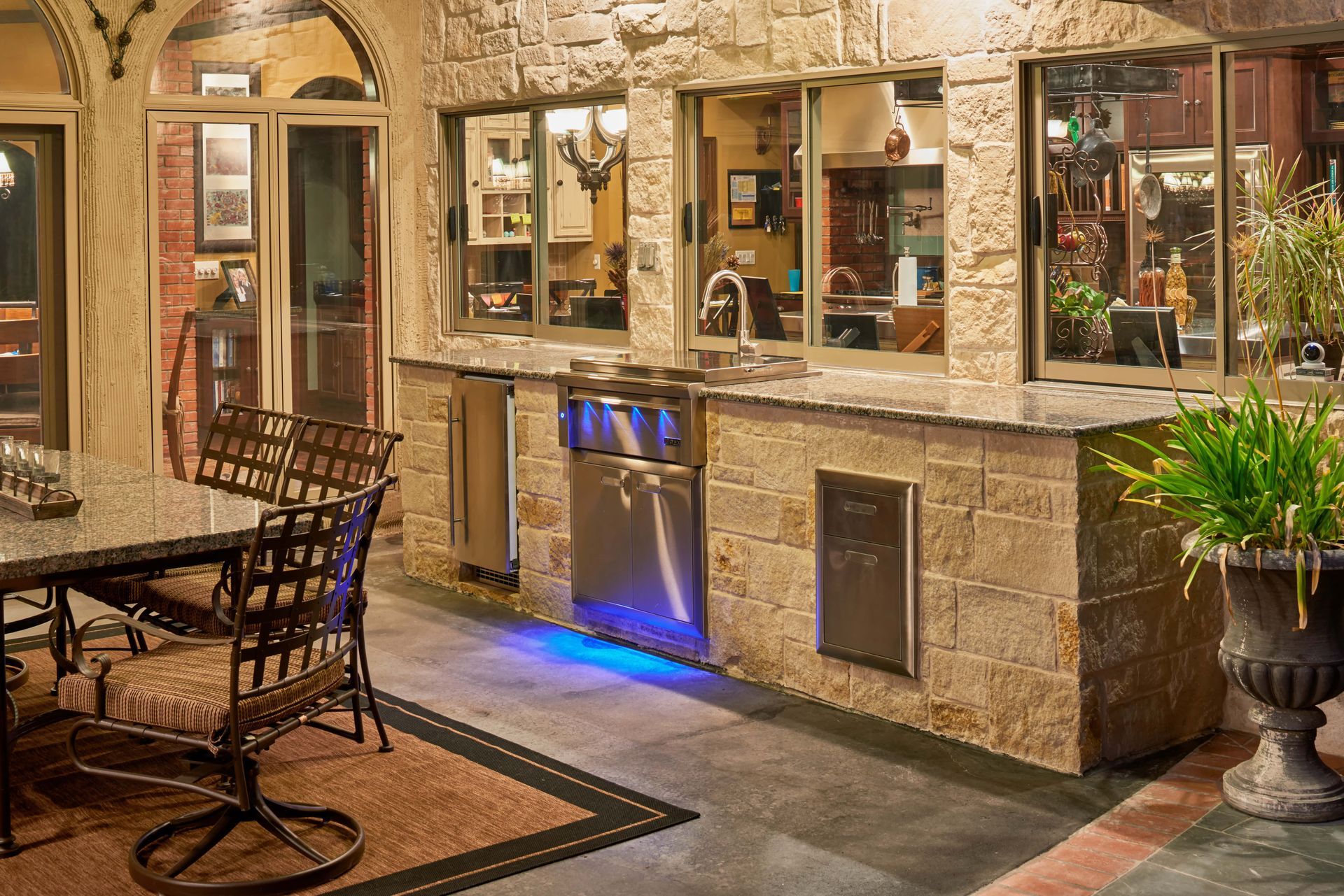 Outdoor kitchen with stone counters, sink, grill, and stainless steel appliances. Dining table nearby.