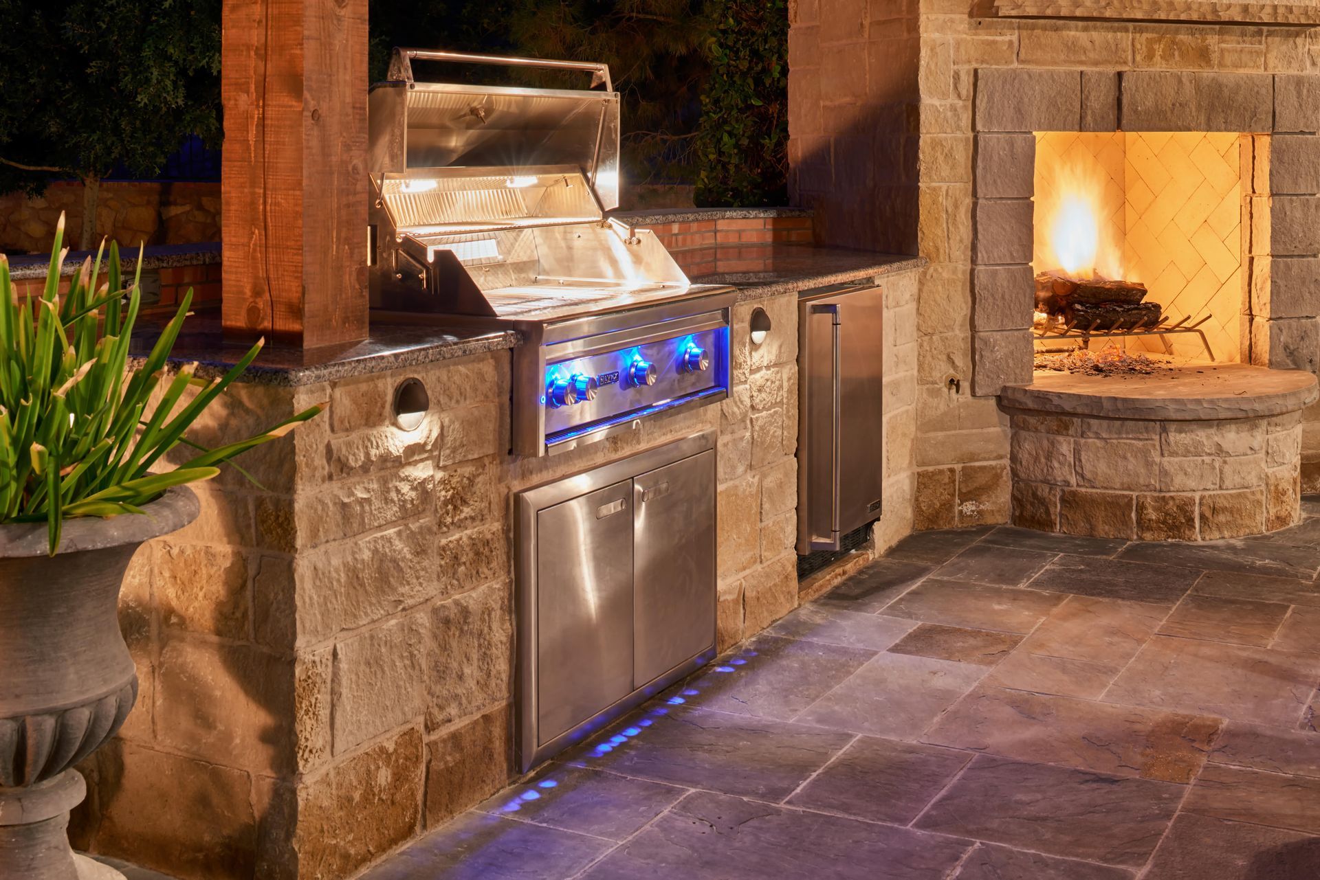 Outdoor kitchen with grill, refrigerator, and fireplace, built into stone structure; warm lighting.
