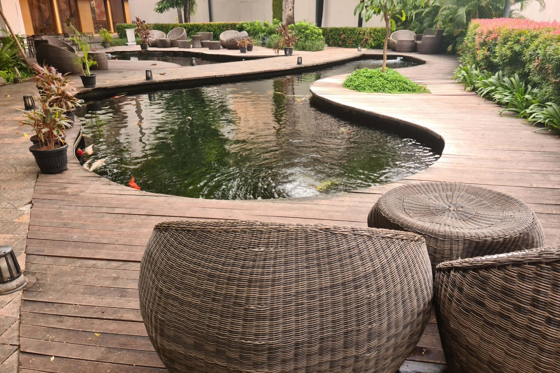 Woven furniture sits near a pond with wooden pathway and landscaping.
