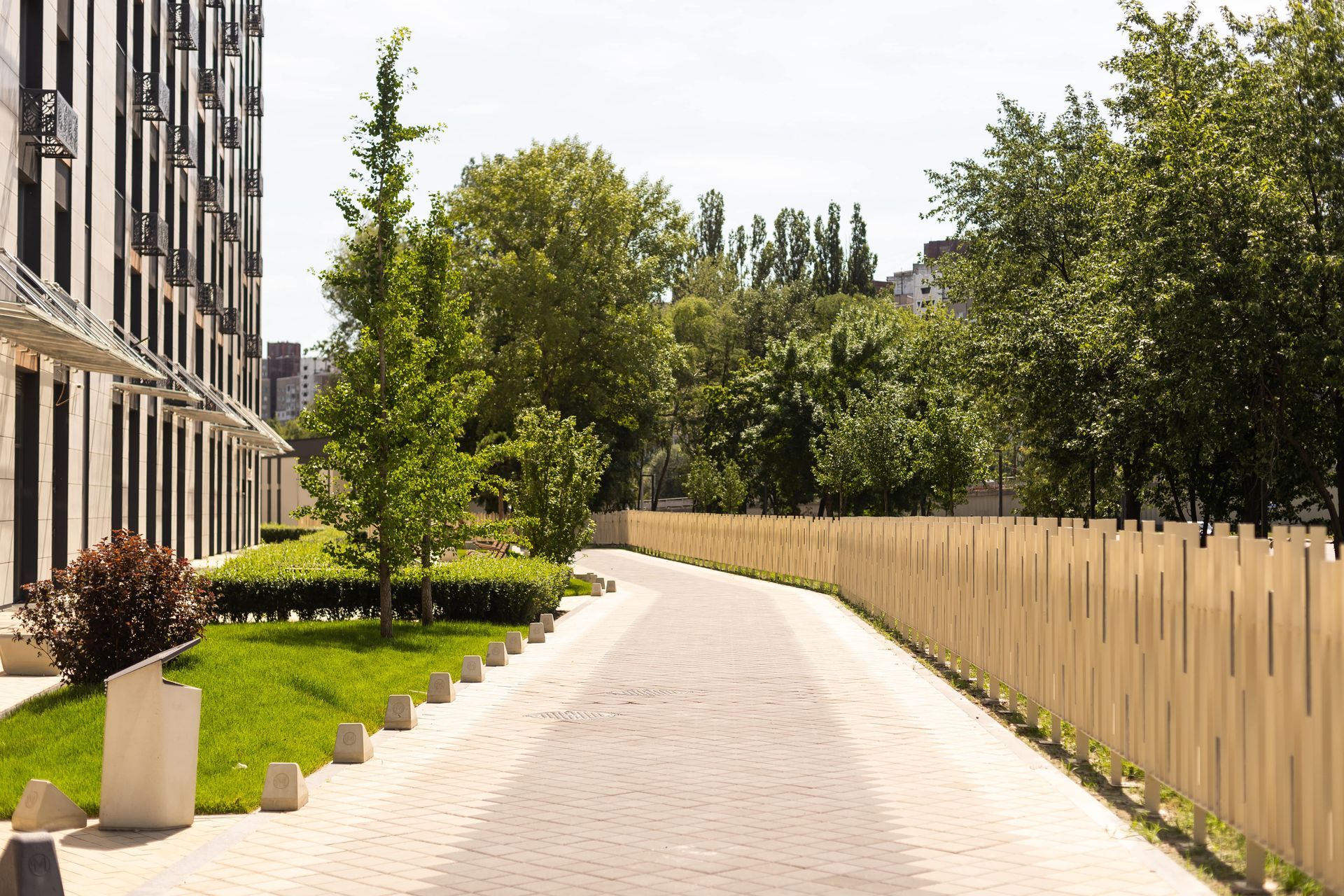 Pathway between building and wooden fence, lined with grass, trees, and planters on a sunny day.
