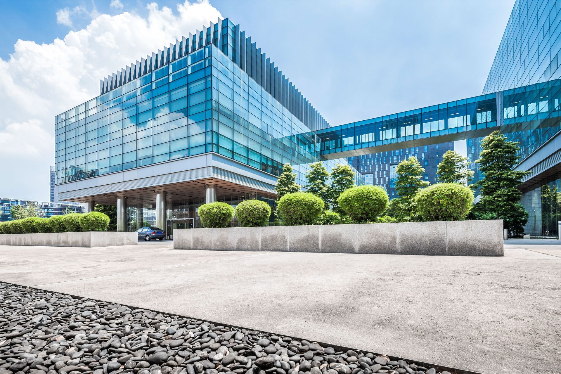 Modern glass and steel office building with a skywalk, blue sky, and landscaping.