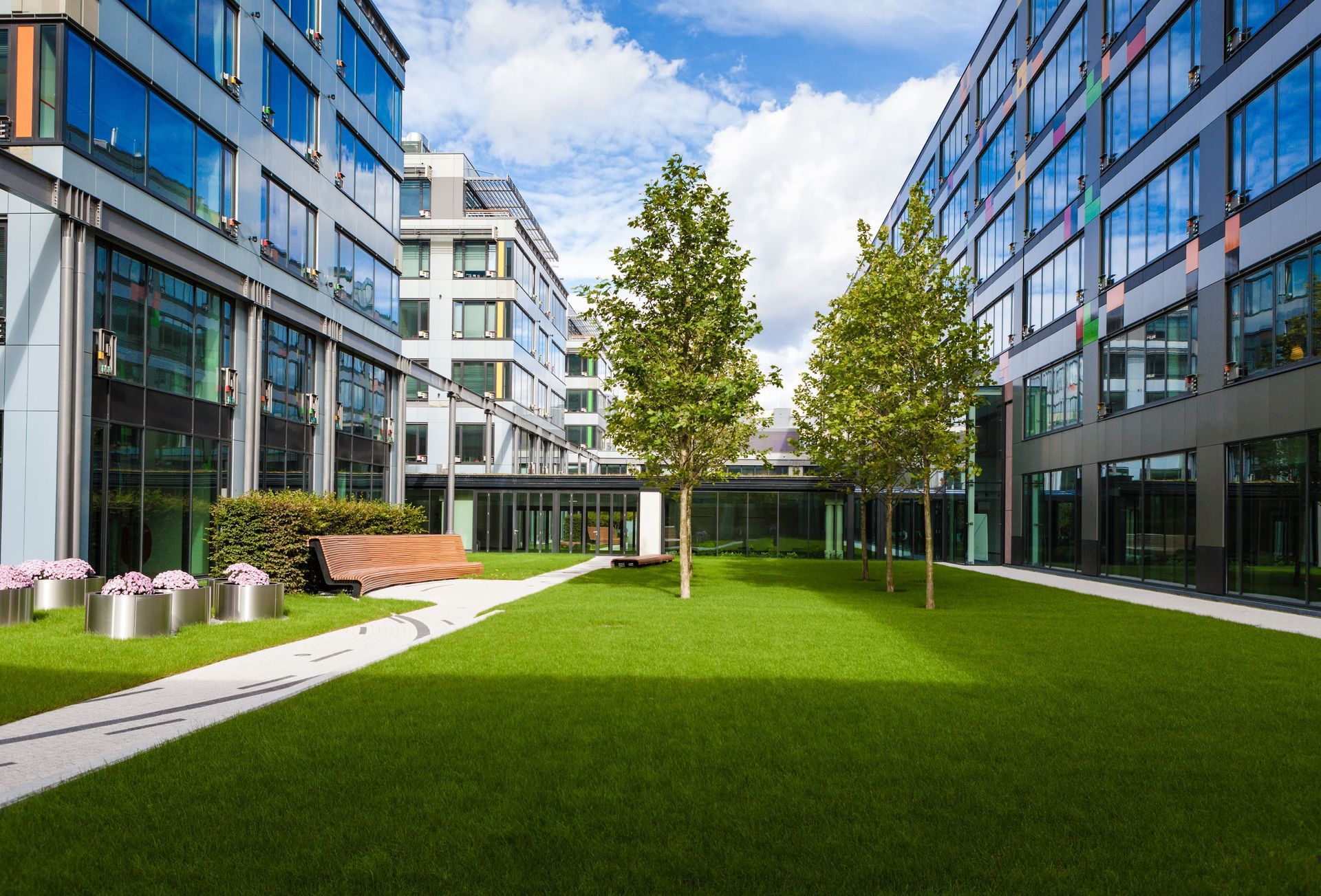 Courtyard with green lawn, trees, benches, and modern buildings. Bright sky with clouds.