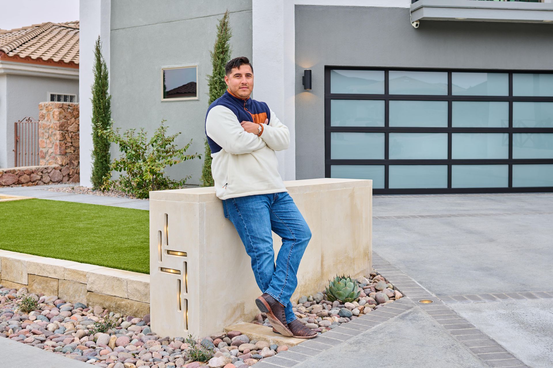 Man leaning on a concrete barrier outside a modern house, arms crossed, wearing a jacket and jeans.