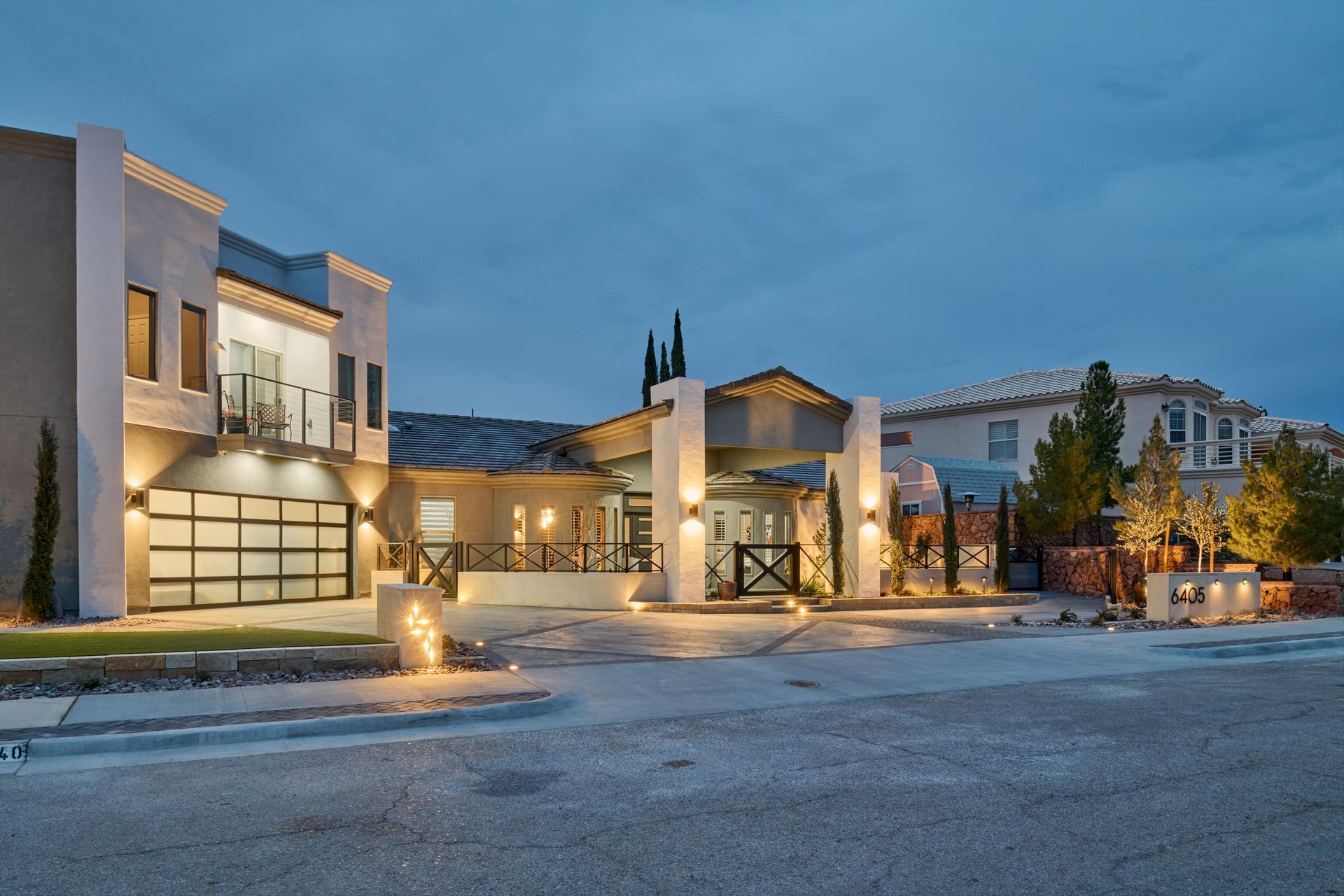 Modern home exterior at dusk, with illuminated facade, garage, and walkway; neighboring homes in the background.