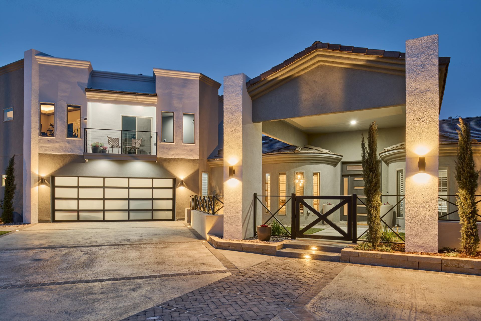 Modern two-story house with illuminated exterior, glass garage door, and decorative pillars at dusk.