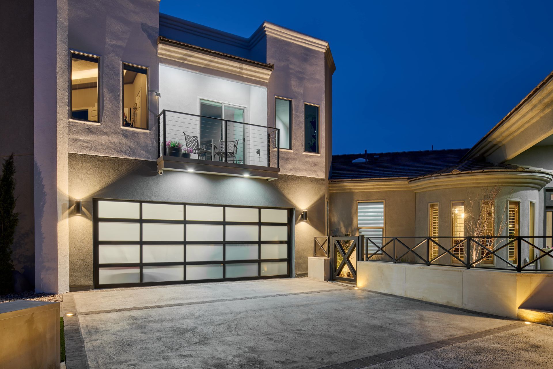 Modern two-story house with a glass garage door and balcony, illuminated at dusk.