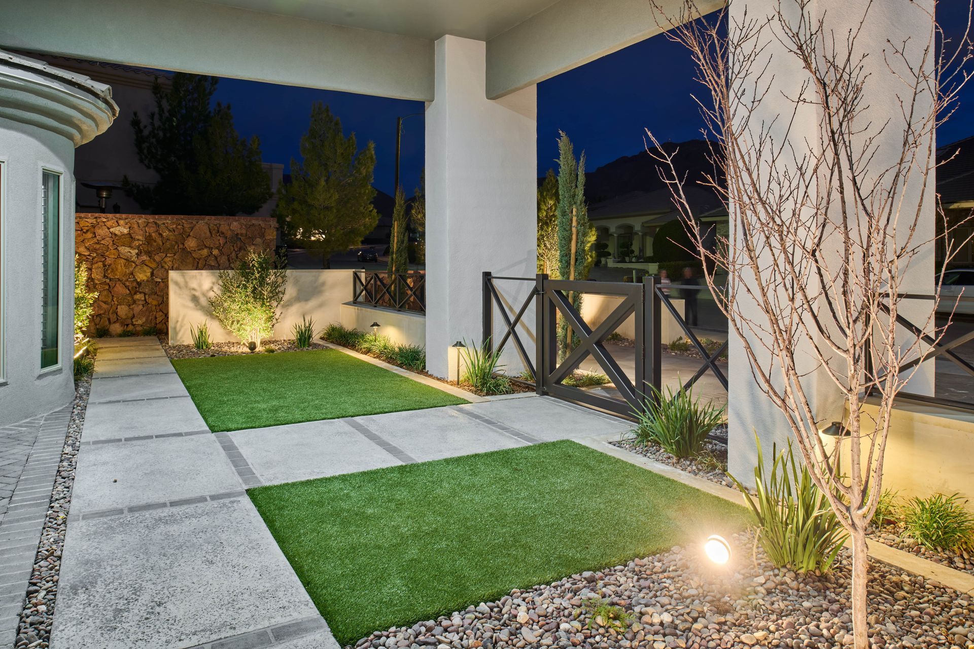Nighttime exterior: Modern patio with grass squares, stone pavers, black gate, and ambient lighting.