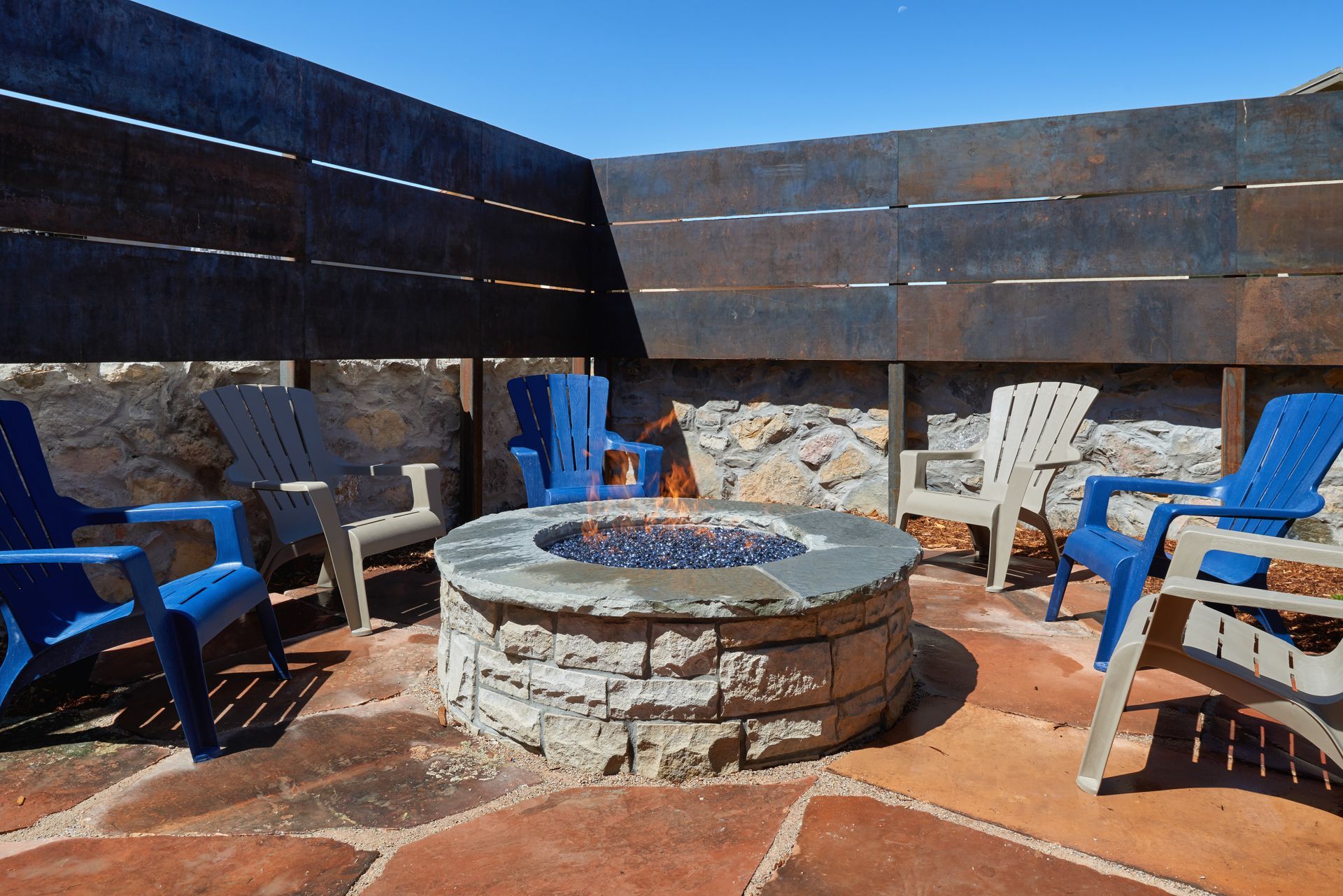 Outdoor fire pit surrounded by blue and beige chairs in a walled patio with a blue sky.