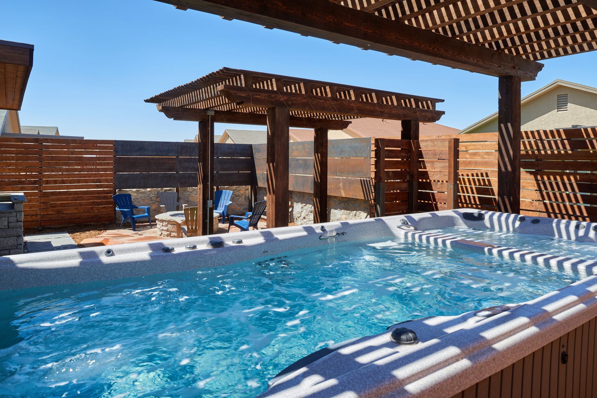 Hot tub under a wooden pergola, with blue water and chairs in the background on a sunny day.