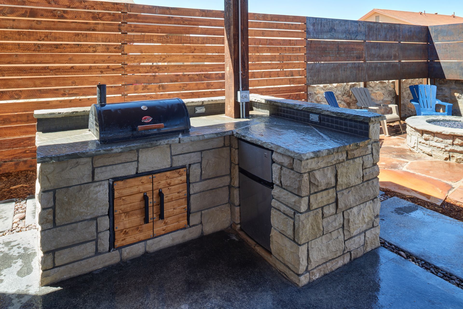 Outdoor kitchen with stone counters, grill, fridge, and wooden cabinets. Wooden fence in background.