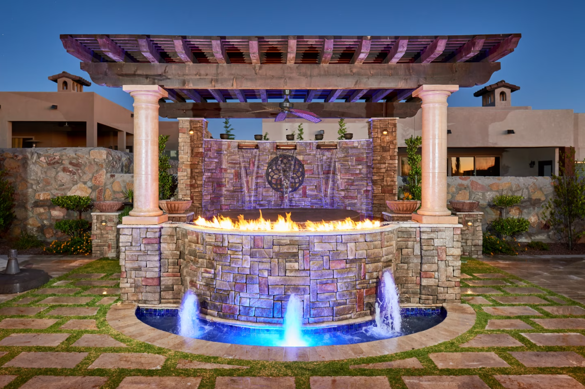 Water feature with a fire pit, fountains, stone wall, and pergola, illuminated with blue and orange lights.