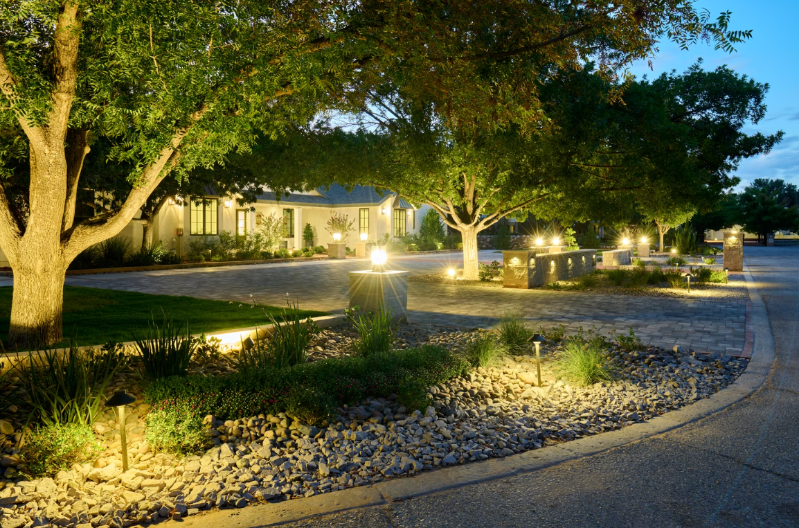 Illuminated trees and landscaping lead to a house on a gravel driveway, with stone and grass accents.